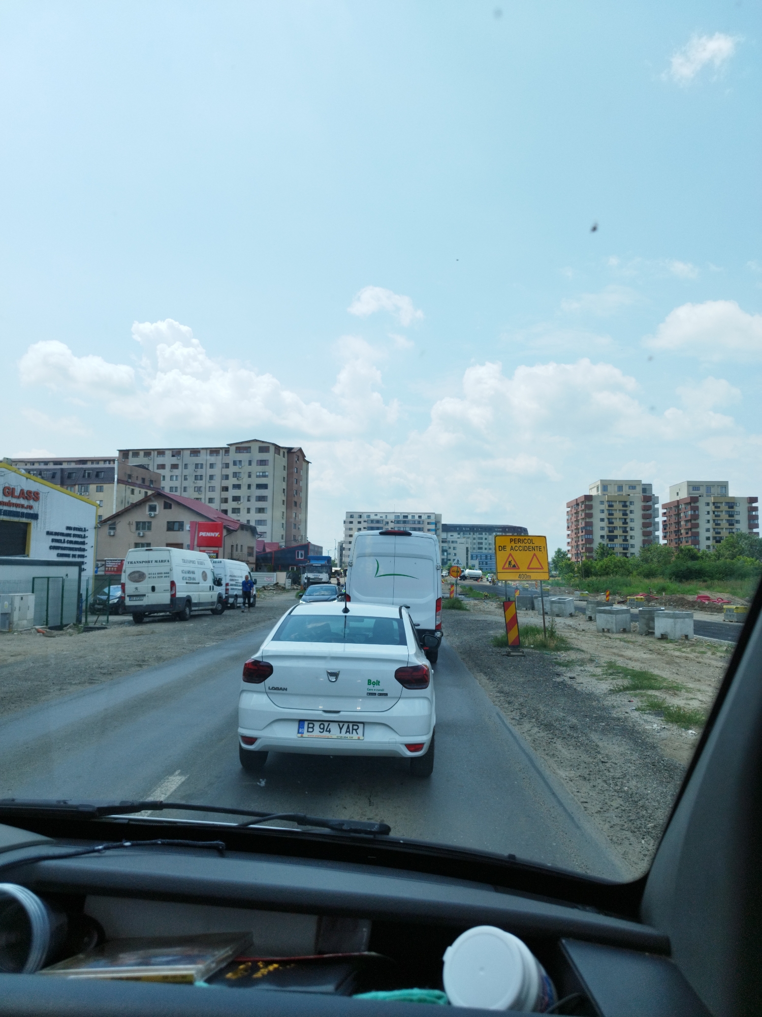 The image captures a street scene in Bragadiru, Romania, likely during daylight hours under a cloudy sky. In the foreground, the interior of a vehicle is visible, including the dashboard and windshield wipers. A white car, identified by its license plate "B 94 YAR", is directly ahead. Further down the road, traffic appears to be moving slowly, with several white vans and other vehicles present.
To the left of the road, a yellow traffic sign with a red border indicates "PERICOL DE ACCIDENTE" (Danger of Accidents) with a warning symbol and "400m" written below. Construction materials, including concrete blocks and red piping, are visible along the roadside, suggesting ongoing roadwork. In the background, multi-story apartment buildings line the horizon. A building with a "PENNY" sign is also visible, suggesting a commercial area. The overall scene depicts a typical urban environment with elements of construction and daily traffic.