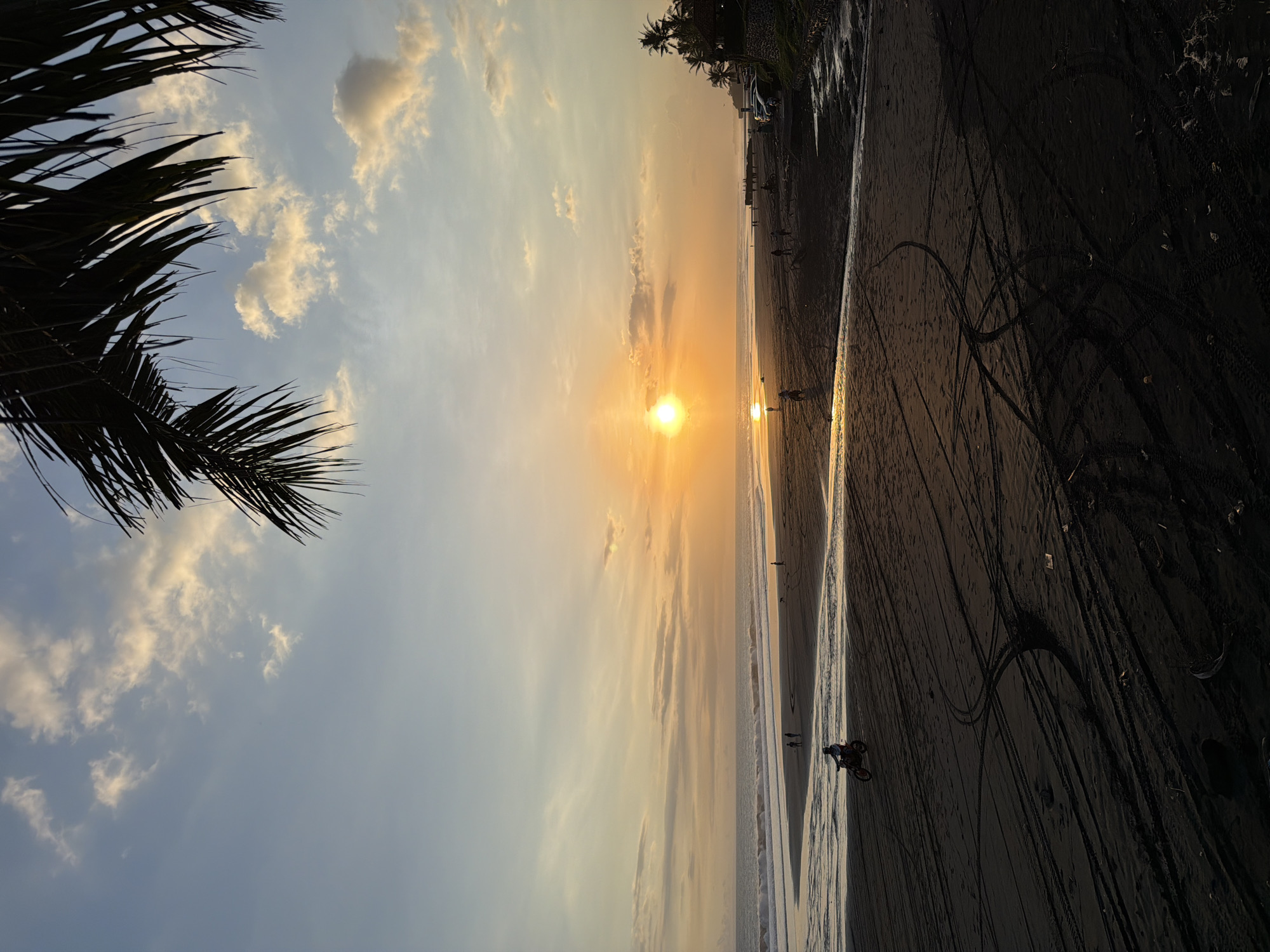 The image shows a sunset at a beach in Kerambitan, Indonesia. The sun is setting over the ocean, casting a golden glow on the sky and the water. Palm trees are visible along the beach. People are walking or riding bikes along the wet sand. The dark sand shows tracks from tires. The sky is a mix of blue and orange colors.