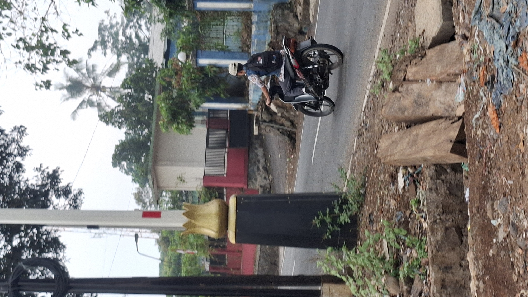 In Cilaku, Indonesia, a person rides a motorcycle on a paved road. The rider, wearing a light-colored cap, a patterned shirt, and dark pants, has a black backpack on. The motorcycle is silver and red. The road is bordered by a rough, debris-strewn embankment with some greenery. A tall, dark-colored pole with a gold, tulip-shaped ornament stands on the left side of the road. Behind the embankment, a building with a red and dark-colored facade and a black metal fence is visible, along with trees and a transmission tower in the background. The sky is overcast.