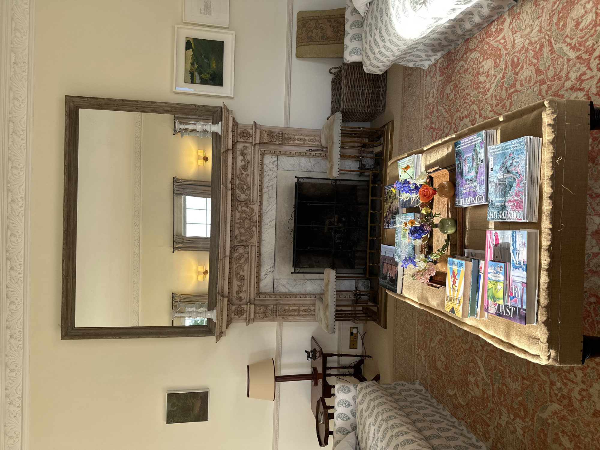 The image shows an interior living space. Prominent in the foreground is a coffee table draped in burlap, holding a variety of books and a floral arrangement. The floral arrangement includes orange roses and blue and white flowers. Behind the coffee table lies an orange and white patterned rug, with parts of a sofa visible on either side, upholstered in a blue and white paisley pattern. At the back, there's a marble fireplace, above which hangs a large wooden-framed mirror reflecting a window. Additional paintings are mounted on the wall, and a lamp sits atop a small table to the right of the fireplace.