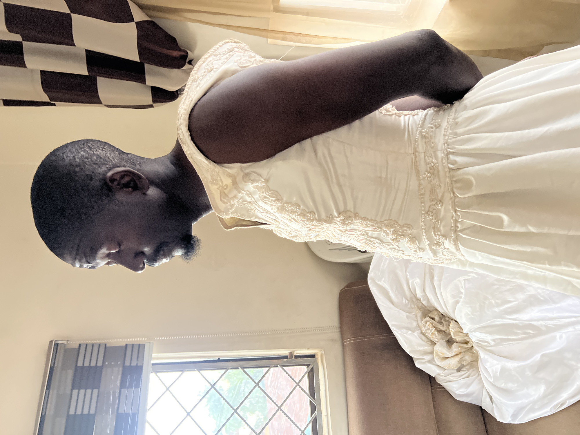 An adult male is standing in a room, facing right, with his eyes closed. He is wearing a white wedding dress that is adorned with beadwork and lace. The dress has a sleeveless design and a full skirt. He has a dark complexion and a short beard. The room appears to be a residential setting, with a window covered by a patterned blind on the left side of the frame. To the right of the window, there is a brown couch. A white garment, possibly another part of the dress or a blanket, is draped over the couch. In the background, a curtain with a checkered pattern of brown and white hangs on the right side of the image. The lighting suggests it is daytime, with natural light coming from the window. The overall environment appears to be a simple indoor space, consistent with a dwelling in Jos, Nigeria.