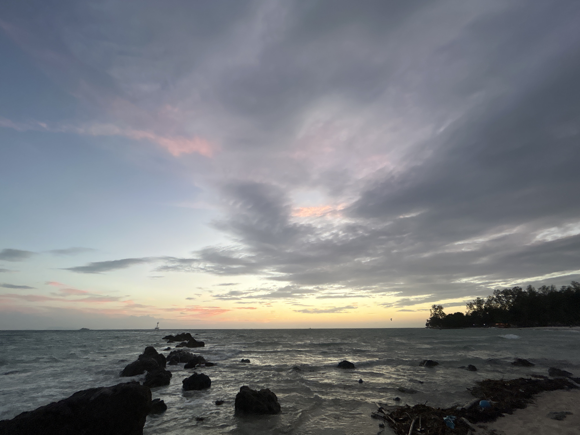 The image depicts a serene beach scene at dusk in Ko Pha-ngan District, Thailand. The sky is a canvas of soft colors, with pastel pinks and oranges blending into blues and grays as clouds drift by. The sea is calm, with gentle waves lapping at the shore. Dark, jagged rocks are scattered along the waterline, some emerging from the water and others resting on the sandy beach. In the distance, the silhouettes of trees line the horizon, and a small boat can be seen on the water. The overall atmosphere is tranquil and picturesque, capturing the beauty of a tropical sunset.