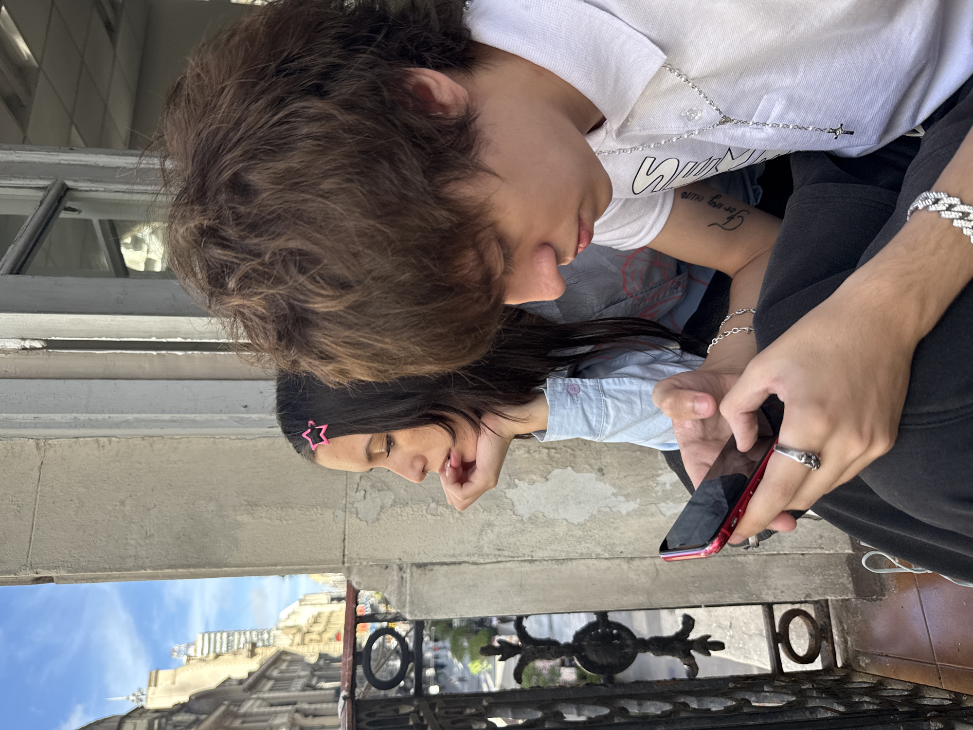 A medium, eye-level, indoor/outdoor shot depicts two young people on a balcony overlooking a street in Buenos Aires, Argentina.
The person in the foreground has dark brown hair and wears a white collared shirt, black pants, and multiple pieces of silver jewelry, including a bracelet, a ring, and a rosary-style necklace. They are looking at a red smartphone held in their hand.
Behind and to the left is another young person with long dark hair, adorned with a pink star-shaped hair clip. They are wearing a light blue button-down shirt. This person is looking in the same direction as the first person and appears to be looking at the phone as well.
The balcony has a concrete wall and an ornate wrought-iron railing. Reflected in the metal railing is a view of the street below, revealing buildings and a blue sky.