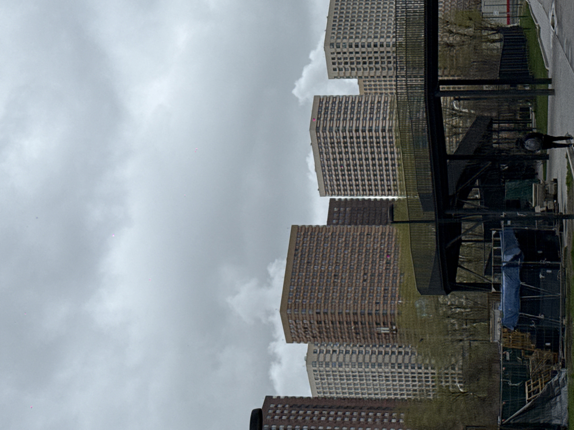 The image displays an urban landscape featuring multiple large, multi-story residential buildings under a heavily overcast, grey sky. The buildings are characterized by their uniform, block-like structure and numerous grid-patterned windows.
On the right side of the frame, a paved pathway or street runs parallel to a dark metal fence. Behind this fence, there are signs of ongoing work or storage, including blue tarpaulins covering materials and various structural elements. A lone individual, dressed in a dark coat and wearing a light-colored hat or hood, is visible walking along the paved path, moving away from the viewer.
The scene conveys a cloudy, possibly cool, daytime environment with no direct sunlight. Small, faint magenta-colored pixel artifacts are scattered across the sky and some building surfaces, appearing to be digital noise rather than features of the scene. There is no visible text. The architecture suggests a contemporary urban setting, consistent with the provided location context of Khimki, Russia.