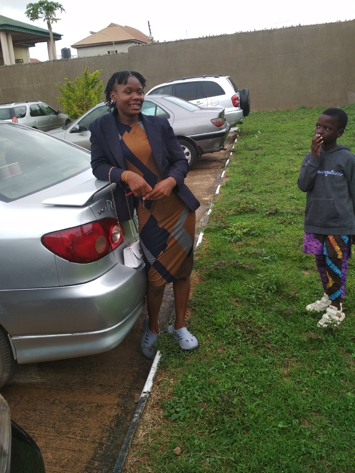 Outside near a silver car in Jos, Nigeria, a young woman in a black blazer and patterned dress leans on the car's trunk, smiling and holding keys with a small white purse dangling. A young boy stands on the grass nearby, wearing a gray hoodie, colorful pants, and white sneakers, with his hand near his mouth. Other vehicles are parked in the background near a tall wall and buildings. The sky appears overcast.