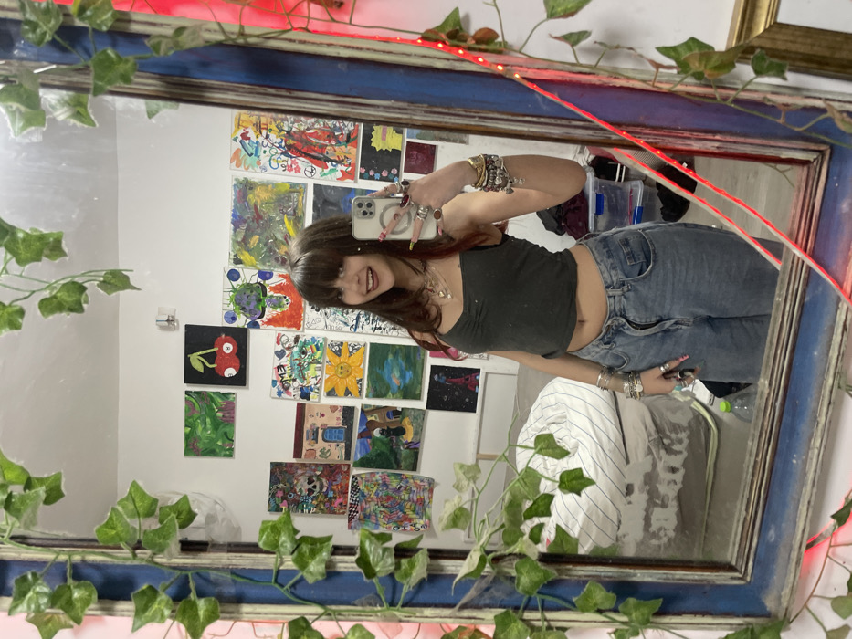 A young woman takes a mirror selfie. She wears a gray crop top and jeans, adorned with rings and bracelets. The mirror, framed in ornate blue wood, is embellished with faux ivy and red string lights. The wall behind her displays an array of colorful, amateur paintings. The room appears to be her bedroom, with a glimpse of a striped futon and storage containers in the background.
