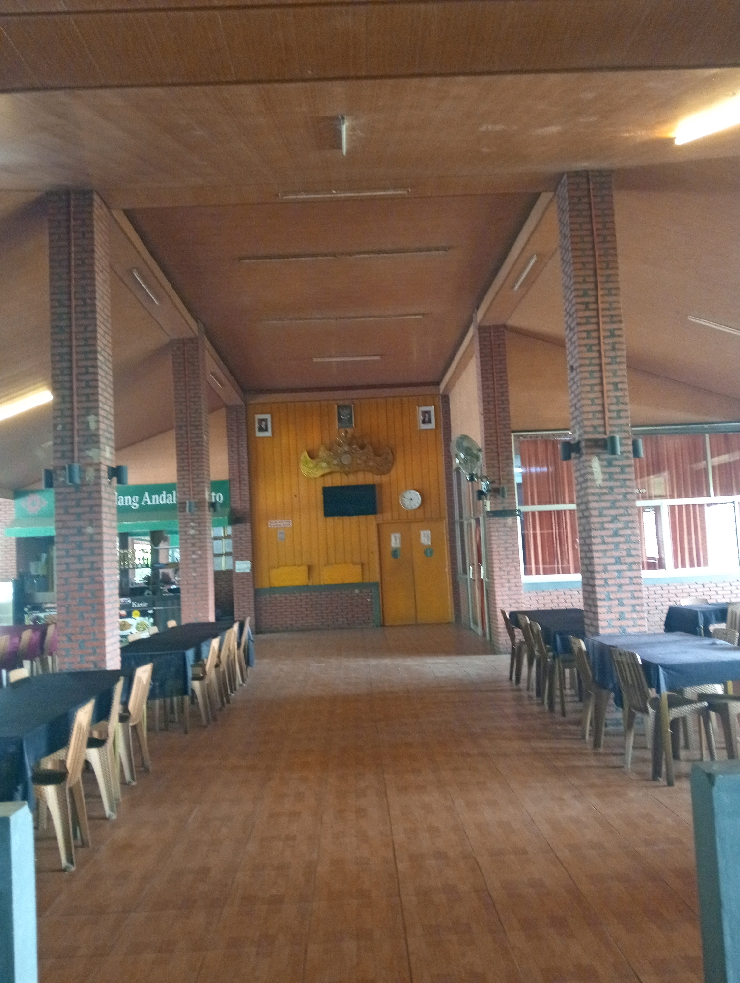 The image depicts the interior of a restaurant or dining hall in Tambak Rejo, Indonesia. The space is characterized by a wooden ceiling with visible beams and paneling, and brick pillars that support the roof. The floor is tiled with a reddish-brown, wood-grain pattern.
The main area features multiple dining tables, most of which are covered with dark blue tablecloths. Chairs, made of a light-colored material, are arranged around the tables. Some tables are set with plates and cutlery. The perspective suggests the photograph was taken from one end of a long hall, looking towards the other.
In the background, a wall is painted yellow and adorned with a decorative golden crown-like ornament and two framed pictures. A television screen is mounted on the wall below the ornament. Two yellow doors are visible on the right side of this background wall, and a white circular clock hangs on the yellow wall to the left of the doors. A green awning with text, possibly a sign for the establishment, is visible on the left side of the image.
On the left side, there is a glimpse of what appears to be a counter or service area, with shelves and possibly food items. A ceiling fan is mounted on one of the brick pillars. Windows with orange curtains are visible on the right side of the background.
No people are clearly visible in the foreground or midground. The environment appears to be an indoor public space, likely a restaurant, designed to accommodate diners.