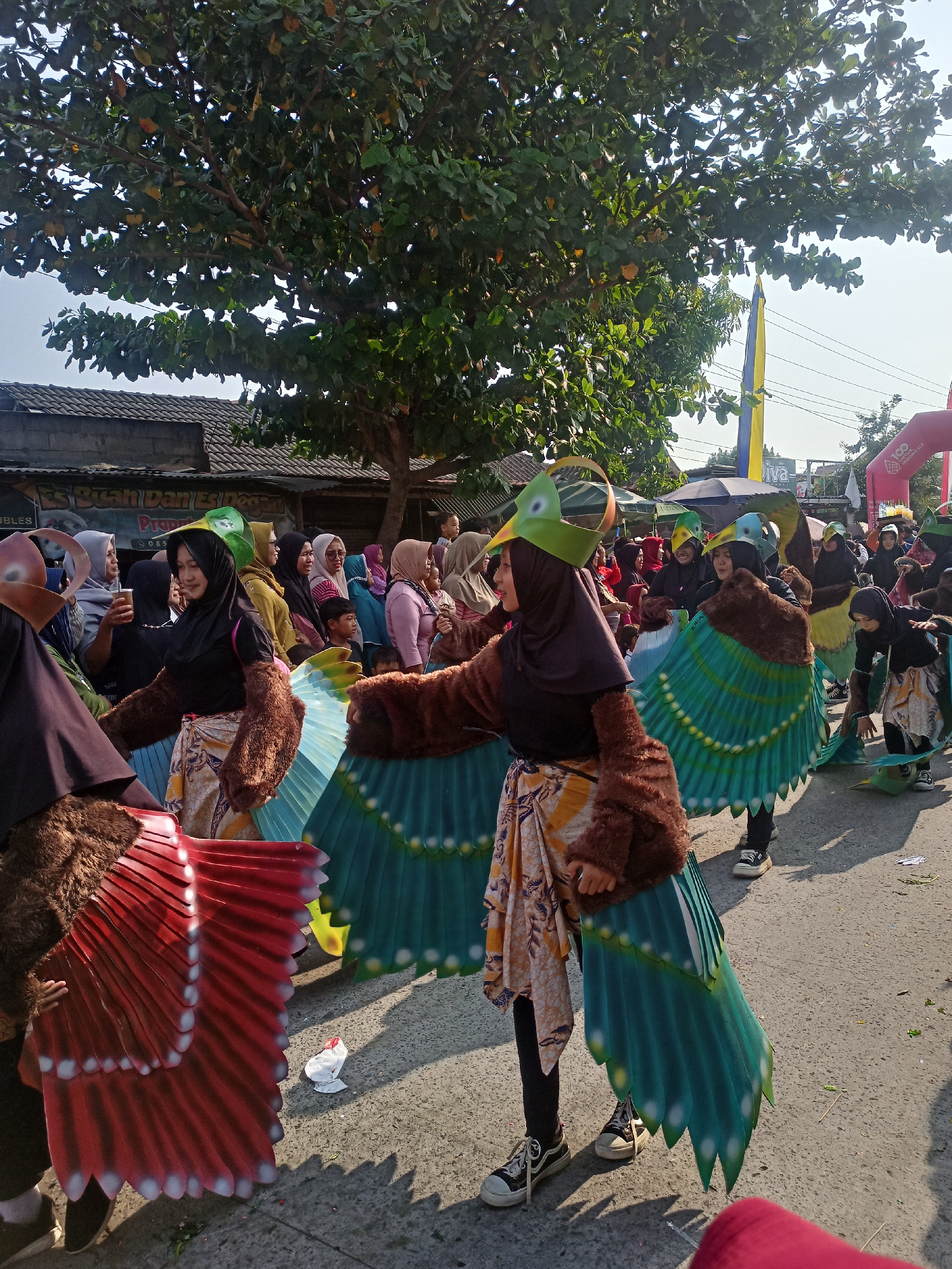 In Saren, Indonesia, a parade is underway featuring participants in elaborate bird costumes. The individuals are adorned with large, colorful wings made of painted cardboard or fabric, designed to mimic bird plumage. Some costumes incorporate brown fuzzy material to represent feathers on the arms and shoulders, while headpieces resemble bird beaks and crests. The participants are walking on a paved road, with a crowd of spectators gathered on the sides, many of whom are also wearing headscarves. In the background, there are buildings with corrugated metal roofs and trees, suggesting a semi-urban or village setting. Banners and a large inflatable structure are visible, indicating a festive event or celebration. The overall atmosphere appears lively and communal, with the parade participants actively engaged in the procession.