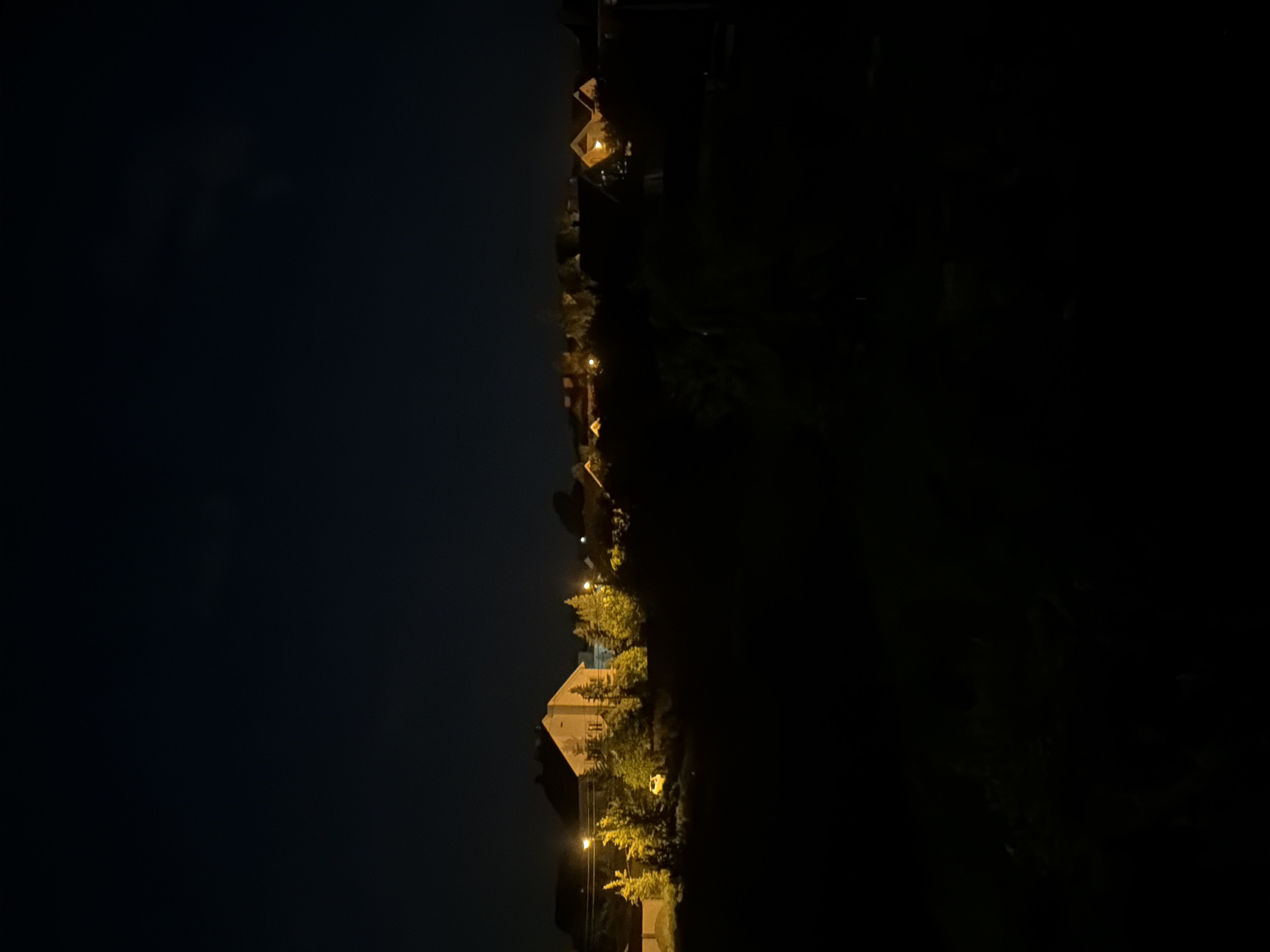 The image shows a night scene in the city of Новинки, Russia. Street lights illuminate a row of houses along a road. The sky is dark, and shadows obscure the areas surrounding the lit road and houses. There are no visible people or active interactions. The primary subjects are the houses, streetlights, and trees along the road. The environment is dark and urban/residential.