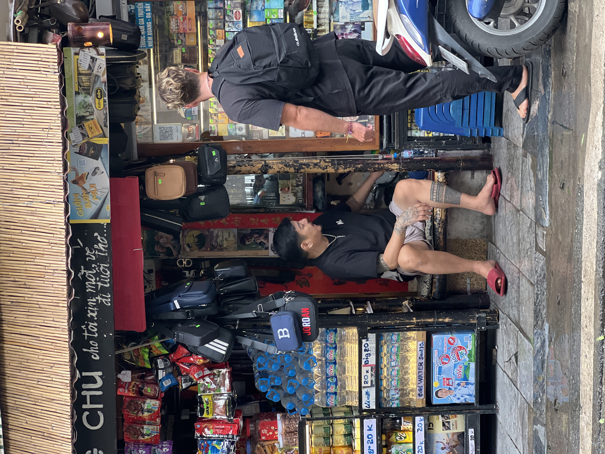 The image depicts a street scene in Hanoi, Vietnam, likely in a commercial area with a small shop. Two men are present. The man on the left, with tattoos on his legs, is sitting on a bench outside the shop, wearing a black t-shirt and light-colored shorts. He is facing the viewer's left. The man on the right is standing, wearing a black t-shirt and black pants, with a black backpack on. He is facing away from the viewer and looking into the shop.
The shop itself appears to be a convenience store or market, with shelves stocked with various goods including water bottles, canned drinks, snacks, and possibly cigarettes. There are several bags hanging from the shop's facade, including backpacks and sling bags. The shop's entrance is partially obscured by the men and displays. A blue scooter is parked to the right of the standing man, with its rear wheel and fender visible. The ground is wet, suggesting recent rain. The background shows more of the shop's interior, with shelves of products. The overall environment suggests a typical urban street scene in Vietnam.