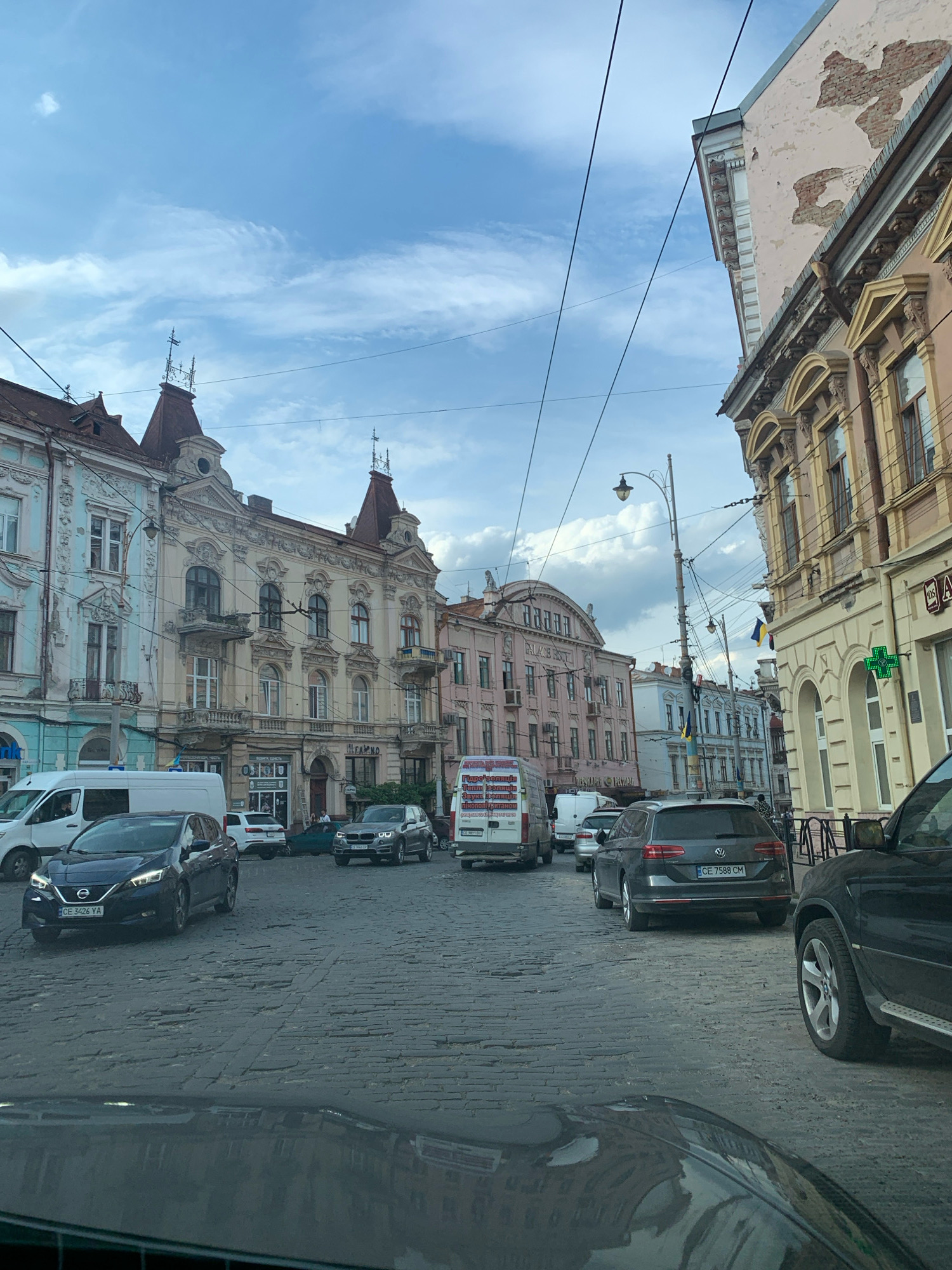 A street scene in Chernivtsi, Ukraine, is captured from a vehicle's perspective, looking forward. The cobblestone street is lined with ornate, old European-style buildings, some with visible architectural details like balconies and decorative facades. Several cars are present on the street, including a dark SUV in the foreground to the right, a dark blue Nissan Leaf in the middle distance on the left, and a grey Volkswagen station wagon further back. A white van is also visible on the left side of the road. Overhead, power lines crisscross the sky, and the sky itself is partly cloudy with patches of blue. Ukrainian flags are visible hanging from some of the buildings. The overall impression is of a historic urban environment.
