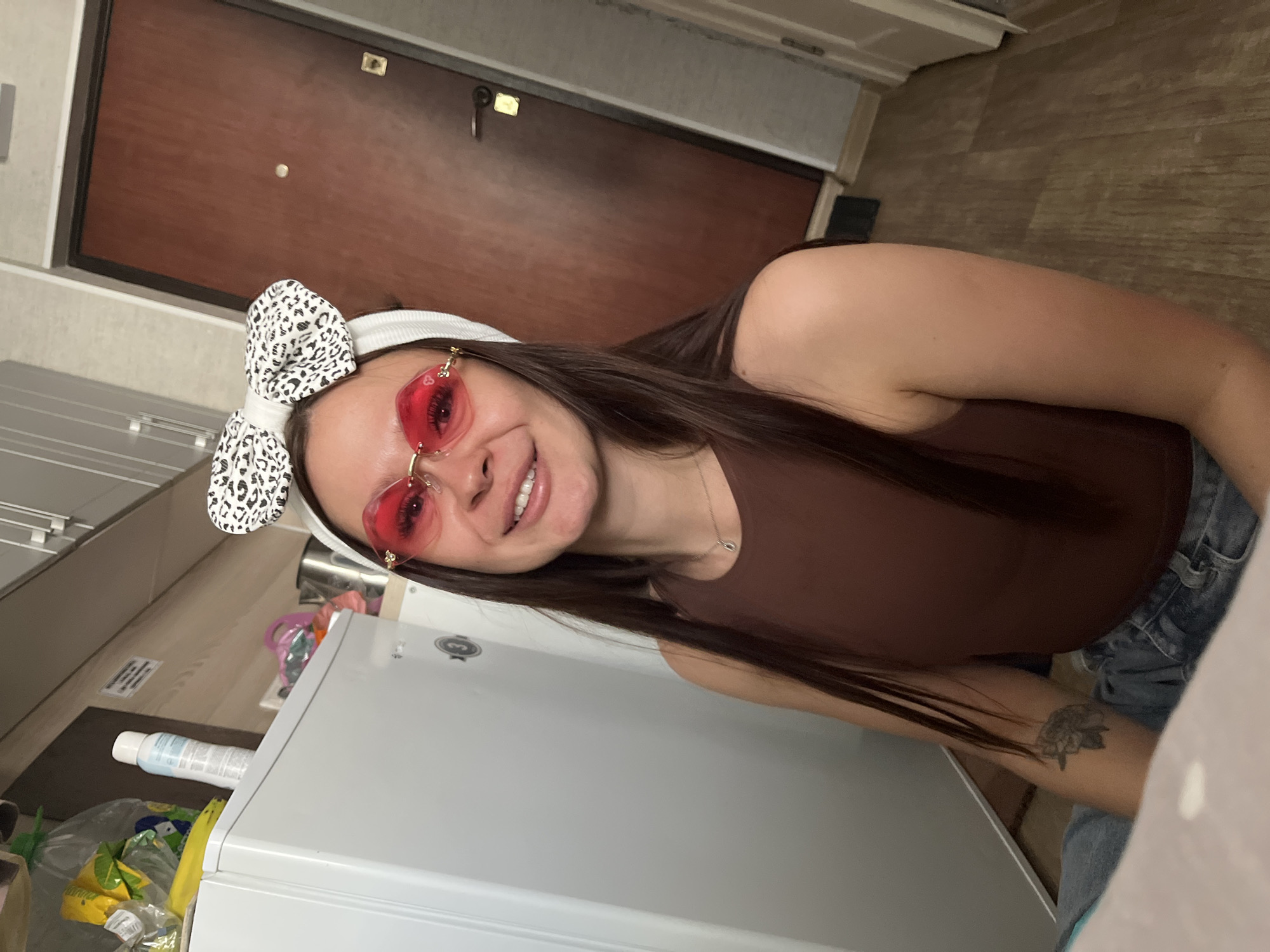 In this indoor shot, a young woman with long, dark hair and a white headband adorned with a black and white leopard-print bow is the central subject. She wears distinctive heart-shaped red sunglasses and a brown tank top. The woman is smiling broadly, her eyes open, suggesting a cheerful or playful mood. Her arm is visible, with a tattoo on her forearm. She is seated near a white appliance, possibly a washing machine or refrigerator, which occupies the lower portion of the frame. In the background, a wooden door is visible, along with part of a hallway. The setting appears to be a domestic interior, likely a kitchen or laundry area. Various objects are scattered around, including a bottle of cleaning product, a plastic bag containing items, and a yellow banana. The lighting suggests it is daytime. The overall impression is of a casual, candid moment captured in a home environment.