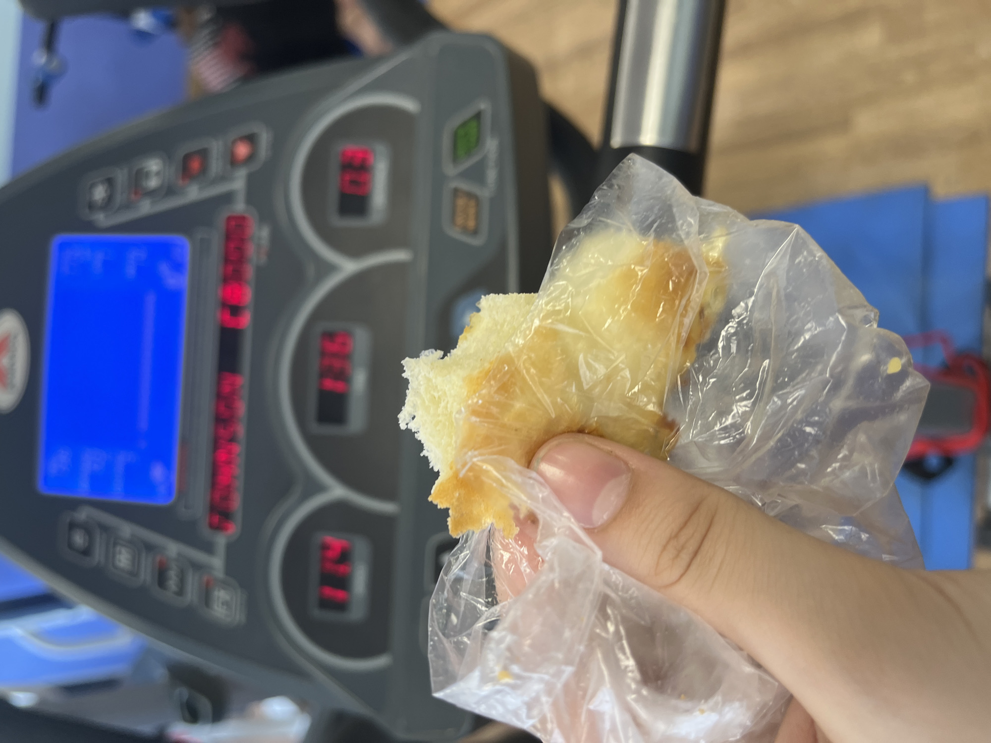 The image shows a partially eaten piece of food, possibly bread or pastry, held in a clear plastic bag by a hand with fair skin. The background is blurred, but it appears to be an exercise machine with a digital display showing metrics, suggesting a gym or fitness center. The display has a blue screen and some numbers in red. The location cannot be confirmed to be Saint Petersburg, Russia, as there is no discernible evidence in the image to support this.