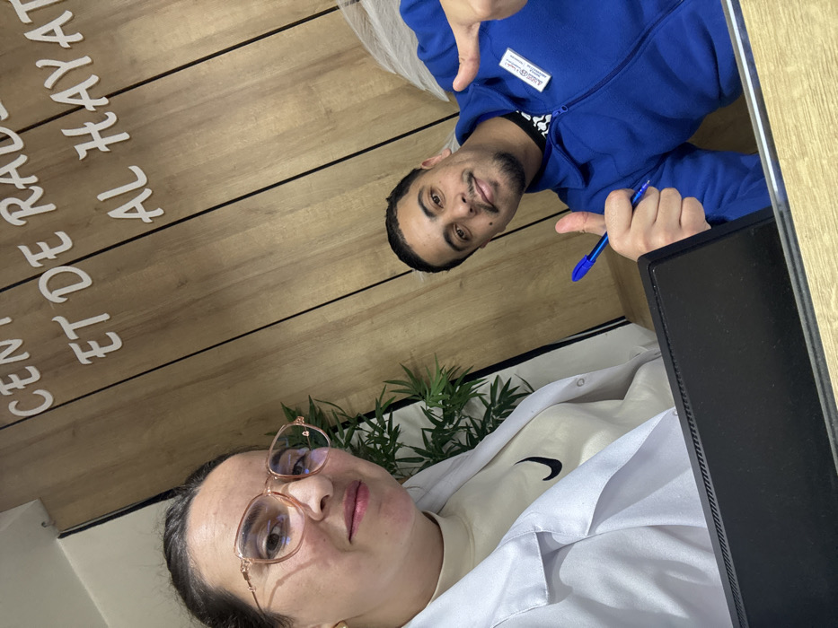 This image captures two individuals, a man and a woman, seemingly taking a selfie or being photographed together indoors. The image is oriented vertically such that the man is positioned towards the upper right and the woman towards the lower left.
The man, who appears to be in his late 20s or early 30s, is positioned in the upper right half of the frame. He has dark, short hair and a neatly trimmed beard and mustache. He is wearing a blue zippered fleece jacket over a dark t-shirt, possibly with a white design (a Nike swoosh is visible on his female companion's shirt, which he might also be wearing underneath). A white name tag or ID badge with text is clipped to the left side of his fleece jacket, near his shoulder. He is looking directly at the camera with a slight smile. His left hand is raised, making a 'thumbs up' gesture, and his right hand is also raised, holding a blue ballpoint pen, while also making a similar 'thumbs up' or pointing gesture with his thumb.
The woman, likely in her 30s or 40s, occupies the lower left portion of the image. She has dark hair pulled back and is wearing a light-colored top (possibly cream or off-white) under a white lab coat or professional jacket. A black Nike swoosh logo is visible on her inner shirt. She is wearing large, gold-rimmed eyeglasses with a slightly pink tint to the frames. She also looks directly at the camera with a neutral to slight smile expression. There's a faint scar or mark visible on her left cheek.
The background primarily consists of a wooden wall or paneling with horizontal planks. On this wooden surface, white raised lettering is visible. The text appears to be in French and partially in Arabic, reading "CENT DE RAD" (partially cropped) followed by "ET DE HAYAT" (partially cropped). The full text likely refers to a "Centre de Radiologie et de Hayat" or similar.
Between the two individuals, towards the center-bottom of the frame, a small, leafy green plant in a pot is visible, adding a touch of decor to the setting. To the right of the woman, a black electronic device, possibly a monitor or part of a computer, is partially visible, suggesting an office or professional environment. The overall impression is that of a bright, well-lit indoor setting, possibly a medical office or reception area, given the lab coat and the visible text.