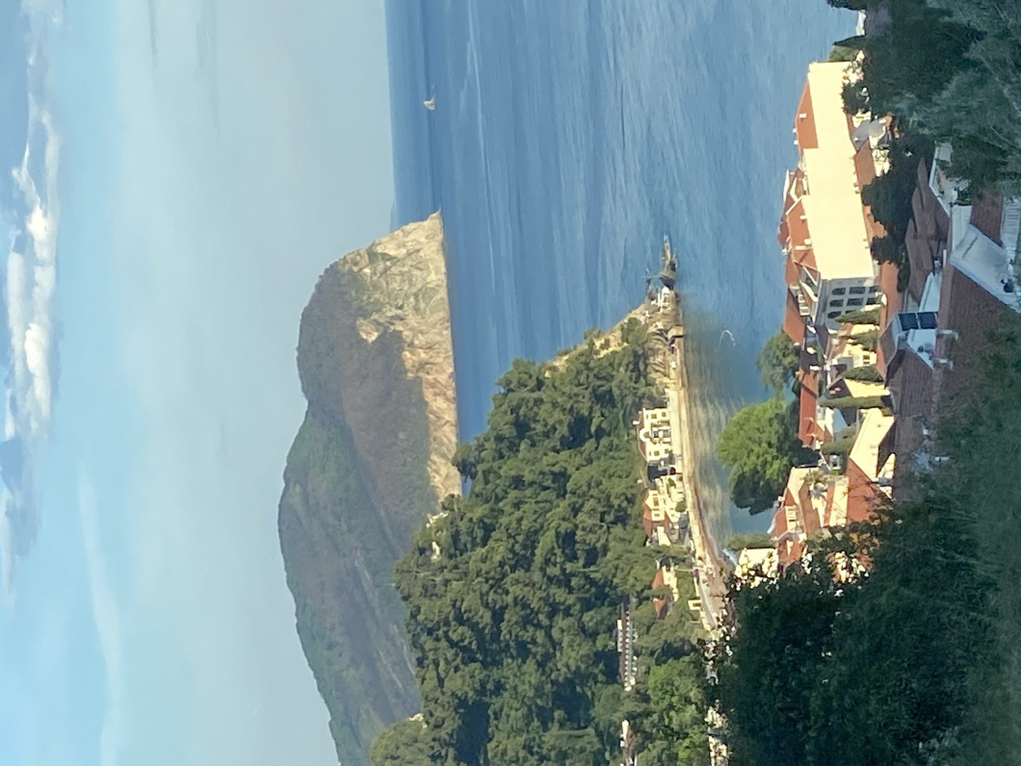 The image depicts a coastal scene in Petrovac na Moru, Montenegro. In the foreground are dense trees with buildings with terracotta roofs beyond. In the middle ground, a rocky peninsula extends into the blue sea with some small buildings along its coast. In the background is a tall, rocky mountain. A sailboat is visible on the water in the distance. The sky is clear with some wisps of clouds.