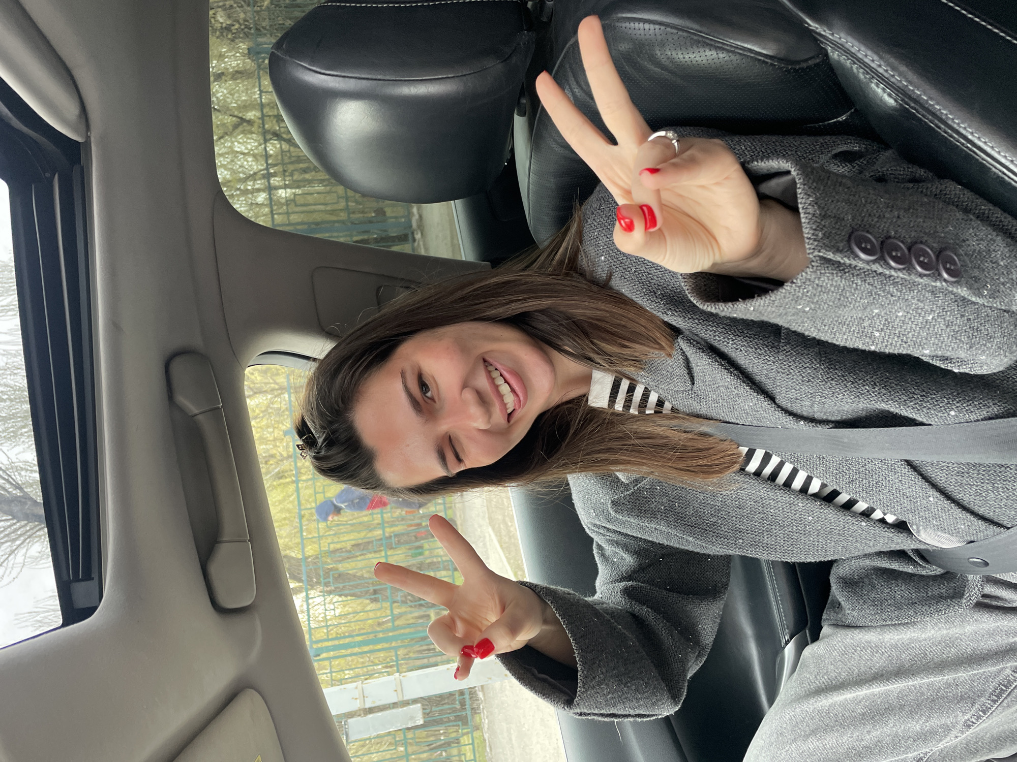 A woman is sitting in the driver's seat of a car, wearing a grey coat, a black and white striped shirt and a seat belt. She is winking with her left eye and holding up the peace sign with both hands, her nails are painted red. The interior of the car is black leather. Outside the window, a fence and some trees are visible, suggesting a residential or park area. Based on the style of architecture and the overall environment, this image could plausibly be located in Krasnoyarsk, Russia.