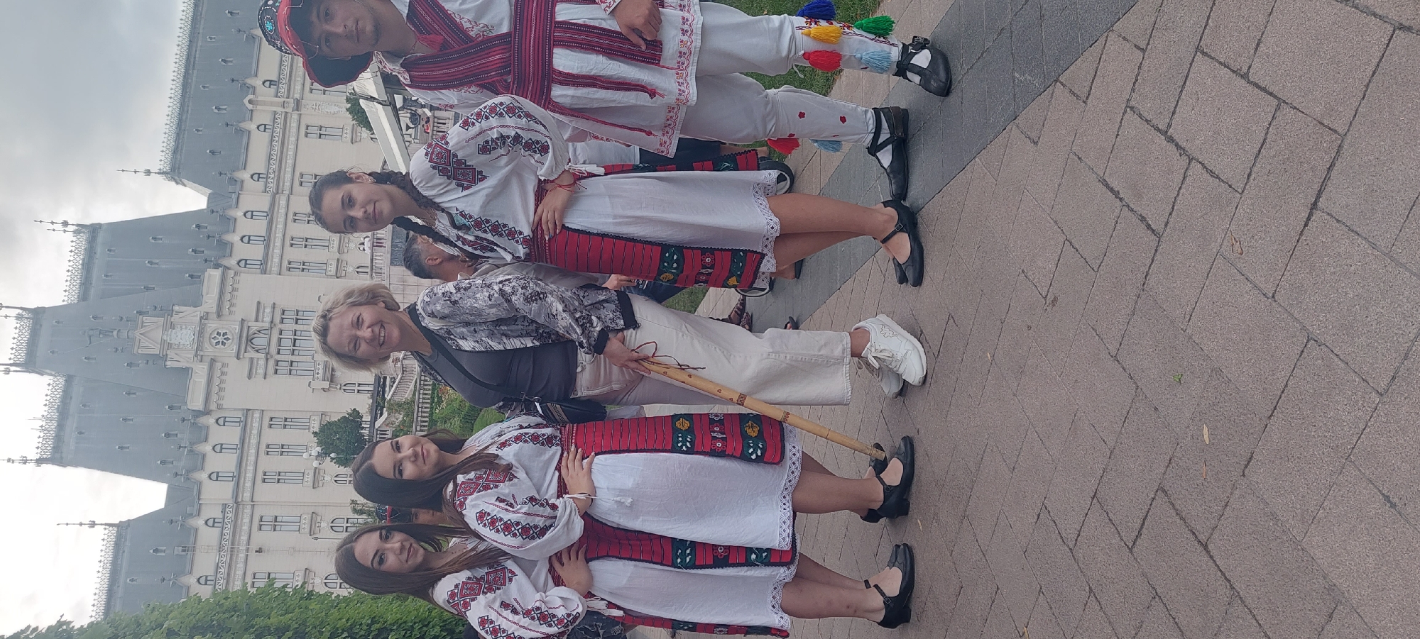 The image shows a group of people in front of the Palace of Culture in Iași, Romania. The Palace of Culture is a grand, imposing building with a distinctive architectural style. In the foreground, a woman is standing with a group of four young people dressed in traditional Romanian folk costumes. The woman is wearing a dark grey t-shirt and light-colored pants, and she is holding a wooden staff. The young people are wearing white blouses with intricate red and black embroidery, and red skirts with matching embroidered sashes. The person on the far right is a young man in a similar costume, including a traditional hat. The people are standing on a paved walkway, and the background shows the impressive façade of the Palace of Culture. The sky is overcast.