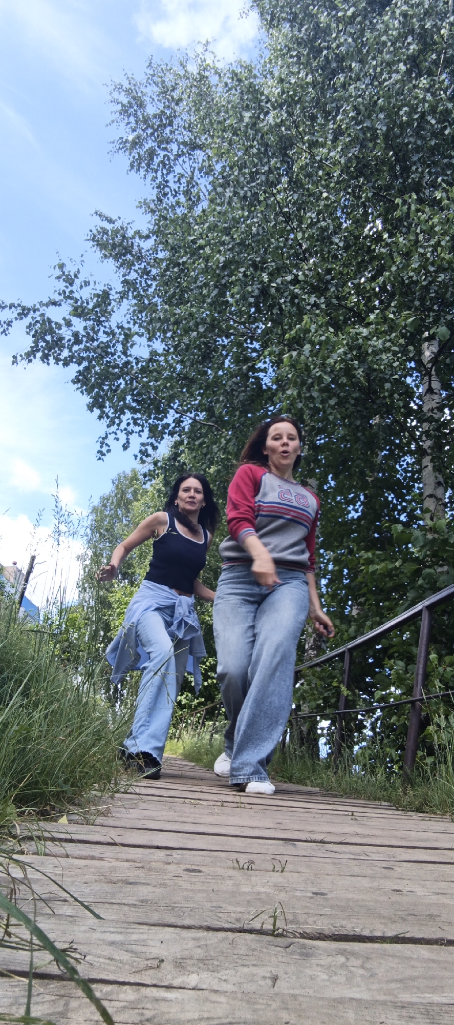 The image depicts two young women descending a wooden walkway. The woman on the left wears a dark tank top, light blue jeans, and a light blue shirt tied around her waist. She has dark hair and is looking forward with a determined expression. The woman on the right is wearing a grey and red long-sleeved shirt and light blue jeans. She has brown hair and appears to be looking slightly ahead and to the side, with her mouth open in a surprised or excited expression. Both women are in motion, suggesting they are running or walking quickly down the path. The walkway is constructed of weathered wooden planks and is bordered by tall green grass on the left and a metal railing on the right. Behind them, a large tree with dense green foliage dominates the background, with a clear blue sky dotted with white clouds visible above. The scene is set outdoors, likely in a park or natural area. The overall impression is one of energetic movement and outdoor recreation. Based on the presence of birch trees and the general landscape, this scene could be located in a natural area within or on the outskirts of Chaikovsky, Russia, a city known for its surrounding forests and proximity to the Kama River.