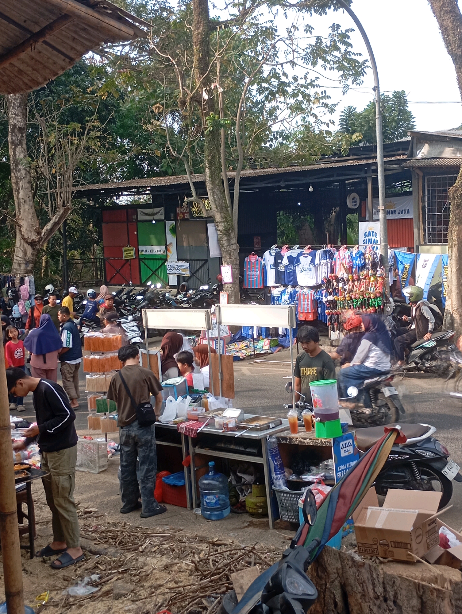 This image depicts an outdoor market scene in Cimahi, Indonesia, likely on a street or open lot, characterized by temporary stalls and a lively atmosphere. In the foreground, a man in a black shirt and olive-green cargo pants stands at a food stall, preparing or serving food. To his right, another man in a brown shirt and camouflage pants is seen from behind, his presence indicating patronage or assistance at a nearby stall. Behind them, a stall displays various goods, including drinks in plastic cups and a large dispenser of purple liquid.
Further back, numerous stalls are set up. One stall features an array of shirts and what appear to be puppets or small figures, arranged for display and sale. Another stall showcases stacked items, possibly merchandise or food supplies, in boxes and containers. Several motorcycles are parked in rows behind the stalls, suggesting a mode of transportation for vendors and customers. A variety of people are visible throughout the scene, browsing, interacting, and going about their day.
The environment is somewhat unkempt, with dry leaves and debris scattered on the ground. Overhead, trees with dense foliage provide shade, and in the background, corrugated metal roofs and simple structures form the market's infrastructure. Banners and signs, some in Indonesian, are displayed on the stalls and buildings, indicating the commercial nature of the location. The overall impression is that of a bustling local marketplace, likely operating during daylight hours.