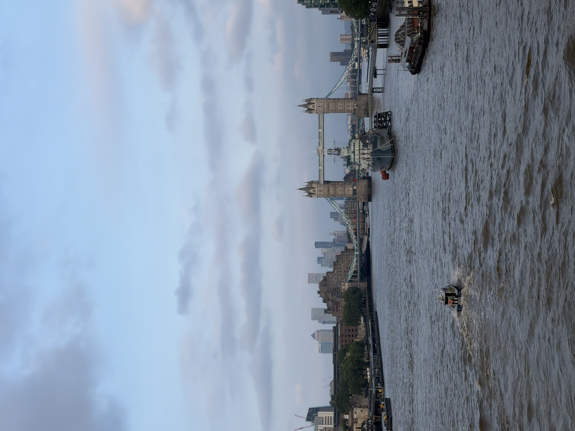 The image shows the River Thames in London, United Kingdom. Tower Bridge is visible in the midground, with buildings on either side of the river. A boat is in the foreground, traveling on the river. The sky is cloudy.