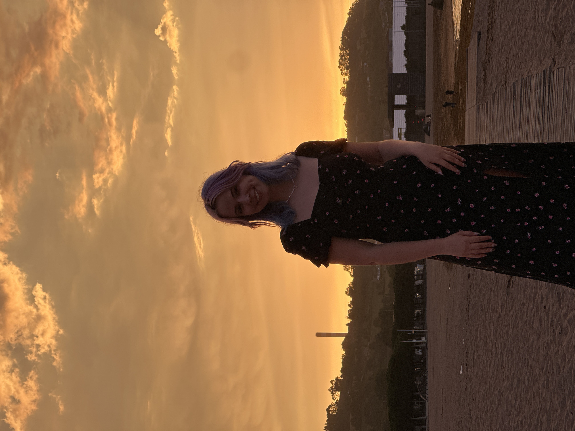 A young woman with purple and blue dyed hair stands on a beach in Gijón, Spain, during sunset. She is wearing a black floral-print dress with a slit on the side and a pearl necklace. The sky is filled with dramatic orange and yellow clouds. In the background, there are trees, a tall industrial chimney, and a body of water. The beach is sandy, with a wooden boardwalk extending towards the water. A few people are visible in the distance on the beach and near the water.