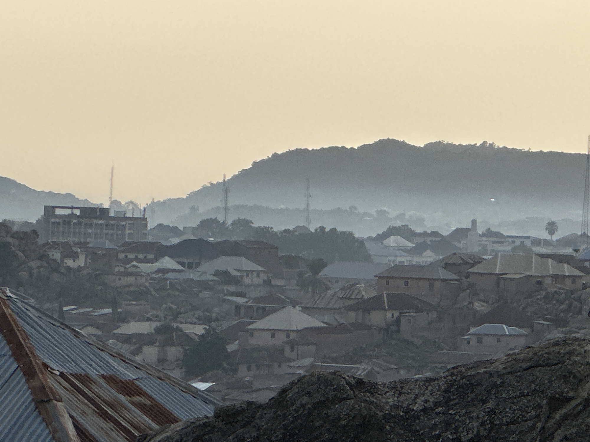 A distant view of a landscape in Jos, Nigeria reveals a densely populated urban area nestled among hills and valleys. The foreground is dominated by a corrugated metal roof and a rocky outcrop. In the mid-ground, a collection of single-story and multi-story buildings with varied roofing materials covers the terrain. Background features include a building under construction, vegetation, and communication towers on the horizon. The sky is bright, indicating early morning hours and limited visibility of the horizon.