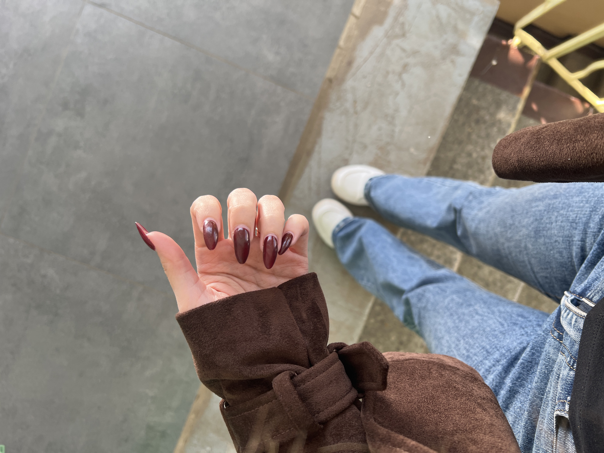 A first-person view captures a hand with long, oval-shaped fingernails painted a deep, wine-red color. The hand is clad in the cuff of a brown suede jacket, adorned with a tied knot detail. The background includes the wearer's legs in blue denim jeans and white sneakers, seated on steps made of gray concrete or stone. A portion of a brown bag rests on the legs. A structure with yellow metal supports is visible behind the steps. The exact location is not discernible from the image, only that it may be in Chaikovsky, Russia.