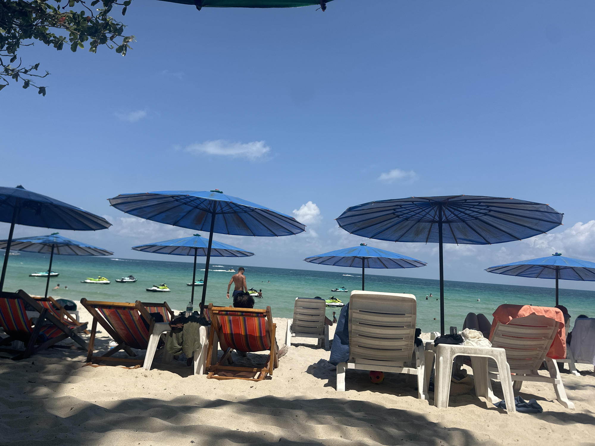 The image shows a sunny beach in Baan Chaweng, Thailand. Several blue beach umbrellas are erected over striped and white plastic beach chairs on a sandy beach. In the background, the turquoise ocean is visible, dotted with jet skis and a few people swimming or wading. A shirtless man is walking out of the water. The scene is partially shaded by trees in the upper left corner.