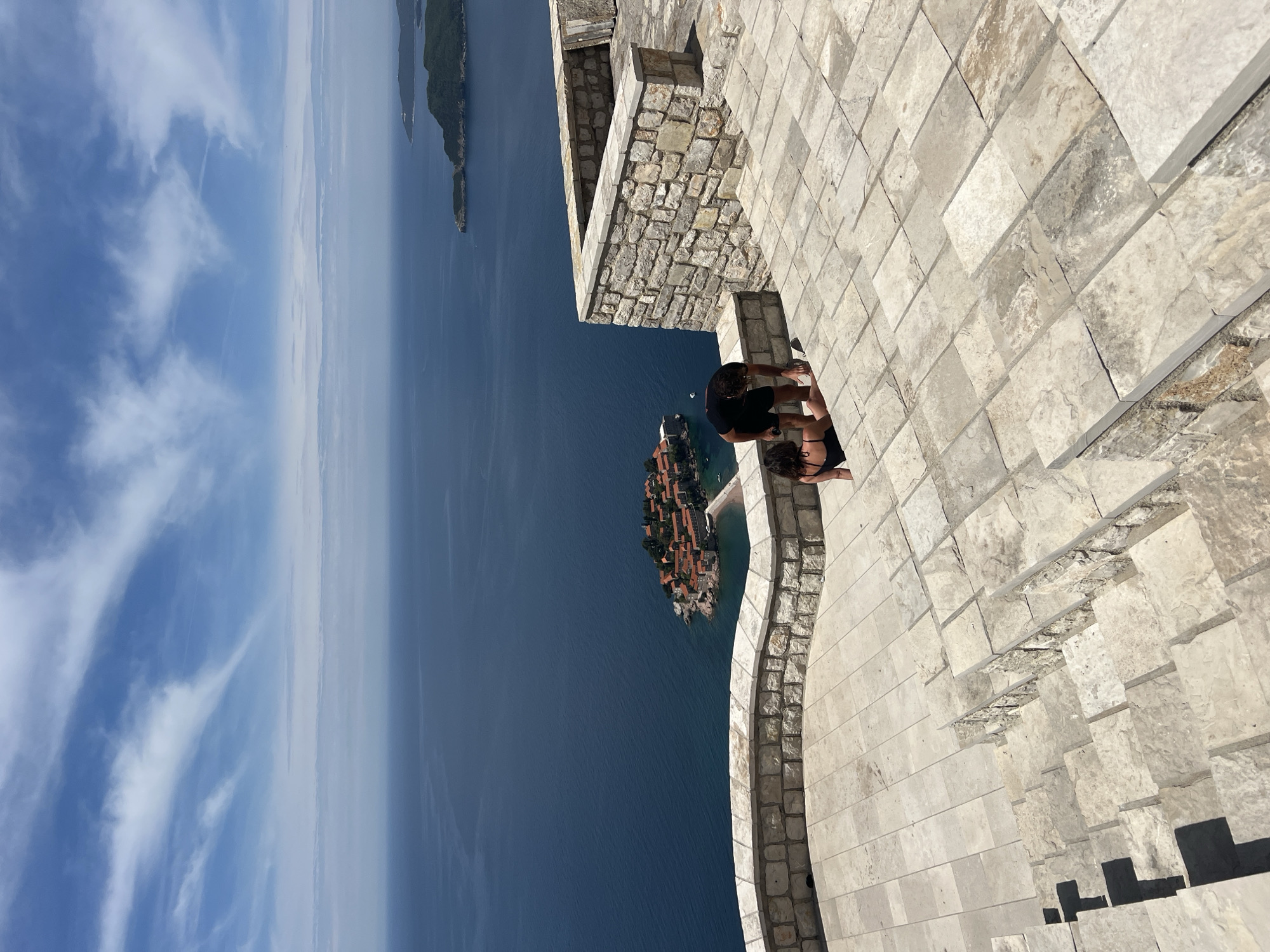 A daytime, medium-distance shot shows a man and a woman on a stone structure, overlooking the Adriatic Sea and the island of Sveti Stefan, Montenegro. The view is from above, looking down and slightly out over the scene.
The couple is near the edge of the stone structure, leaning slightly over the ledge, appearing to look at the island. The man is wearing a dark t-shirt. The woman is wearing a dark bathing suit.
In the background is the Adriatic Sea, with the island of Sveti Stefan visible in the center. The sky is visible in the distance, with clouds and a bright blue color.
The structure the couple is on appears to be a wall or tower, constructed of light-colored stone bricks. The structure includes steps and a lower stone wall. The image is taken from slightly above the couple, emphasizing the height and the drop to the sea below.