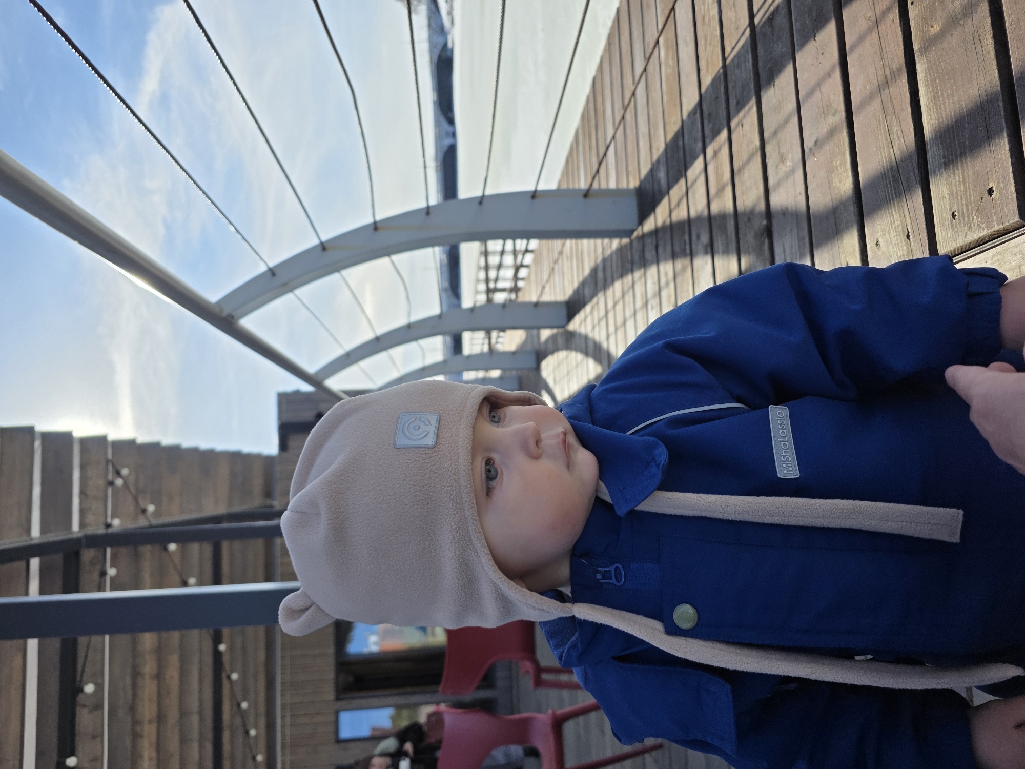 A young child, approximately a toddler, stands in the foreground on a wooden pier in Votkinsk, Russia, under bright daylight. The child wears a beige fleece hat with small ear-like protrusions and a dark blue winter jacket with a "MishaLassie" brand tag visible on the left chest. The child's gaze is directed towards the right. A hand, presumably an adult's, is partially visible at the bottom right, lightly contacting the child's left arm.
The pier features light brown wooden planks, casting distinct, elongated shadows to the right. A light-colored, arched metal railing with multiple horizontal tensioned cables runs along the right side of the pier, separating it from a vast expanse of frozen, snow-covered water. In the far distance across the frozen water, a multi-arch bridge is visible. To the left of the child, a modern building constructed of horizontal wooden planks is partially visible, featuring large windows. Below its overhang, a string of bare light bulbs is suspended. Two red chairs and the partial figure of another individual are present near the building on the left. The sky above is clear blue with minimal cloud cover.