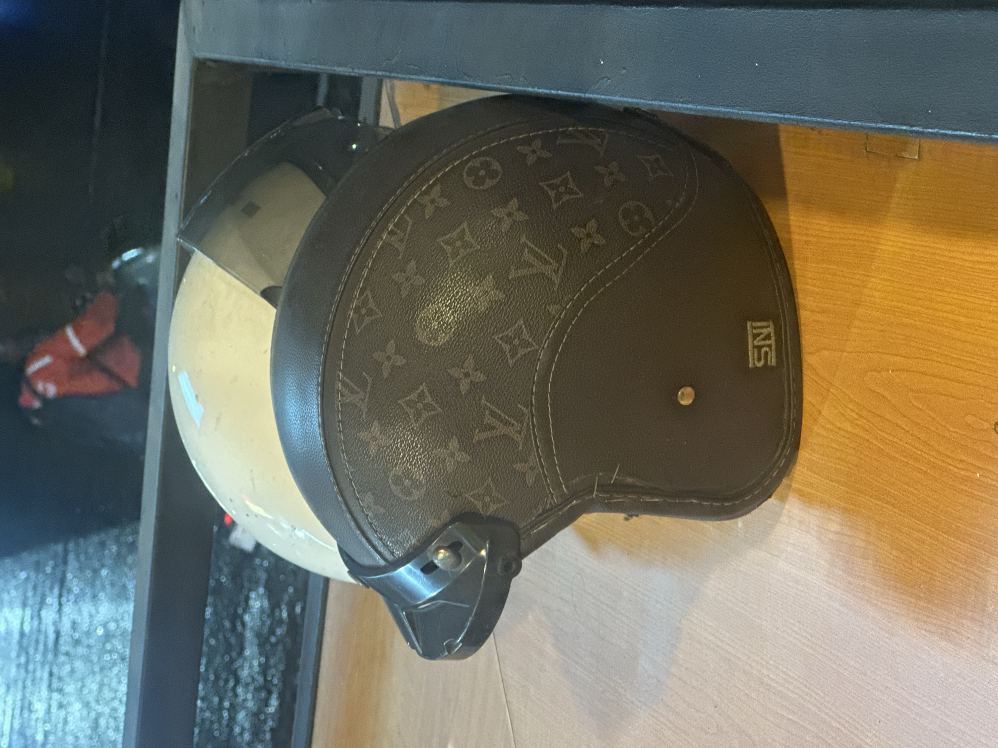 This is a close-up, eye-level shot of two helmets resting on a wooden surface, possibly a shelf or table, with a dark metallic bar overhead. The helmet in the foreground is dark brown with a repeating pattern of the Louis Vuitton monogram. The other helmet, partially visible behind it, appears to be cream-colored with a clear visor. The background on the left is dark and out of focus, with some reflective elements suggesting a wet or shiny surface. The right side of the image shows a light-colored wooden floor with some shadows.
A small, square label with the text "SNI" is visible on the side of the patterned helmet, which is likely a certification mark. The helmets appear to be stationary. There are no people or animals visible, and the lighting suggests it could be indoors or outdoors in low light conditions, possibly evening. The overall impression is one of objects stored or displayed.