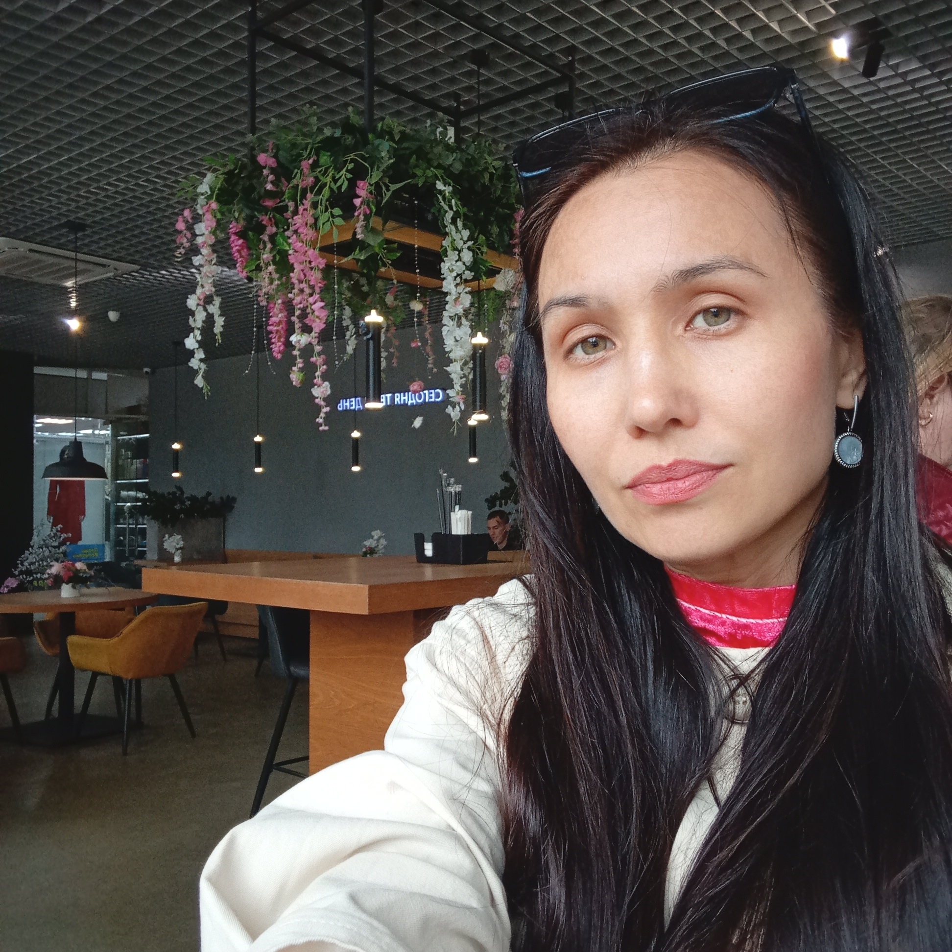 A woman with long, dark hair and light green eyes is captured in a selfie, positioned indoors at what appears to be a cafe or restaurant in Pervomaysky, Russia. She is wearing a light-colored, possibly cream-colored, top with a red high-collared garment underneath, and a circular, light-blue gemstone earring is visible on her right ear. Sunglasses are perched atop her head.
The background features a dark, gridded ceiling with various light fixtures. Hanging floral decorations, primarily pink and white wisteria-like flowers mixed with green foliage, are suspended from the ceiling. Multiple wooden tables and chairs, some in a yellow-mustard upholstery and others dark-colored, are arranged in the open space. In the mid-ground, a black table caddy holding napkins and utensils is visible on a table. A man with dark hair is partially visible sitting at a table further back on the right.
A reflective surface in the background reveals the inverted Russian text "СЕГОДНЯ ДЕНЬ" (SEGONYA DEN', meaning "TODAY IS THE DAY"), and to the far left, a reflection shows a red dress hanging, possibly within an adjacent retail space. The floor is a dark, uniform color. The overall environment suggests a modern casual dining or lounge setting.