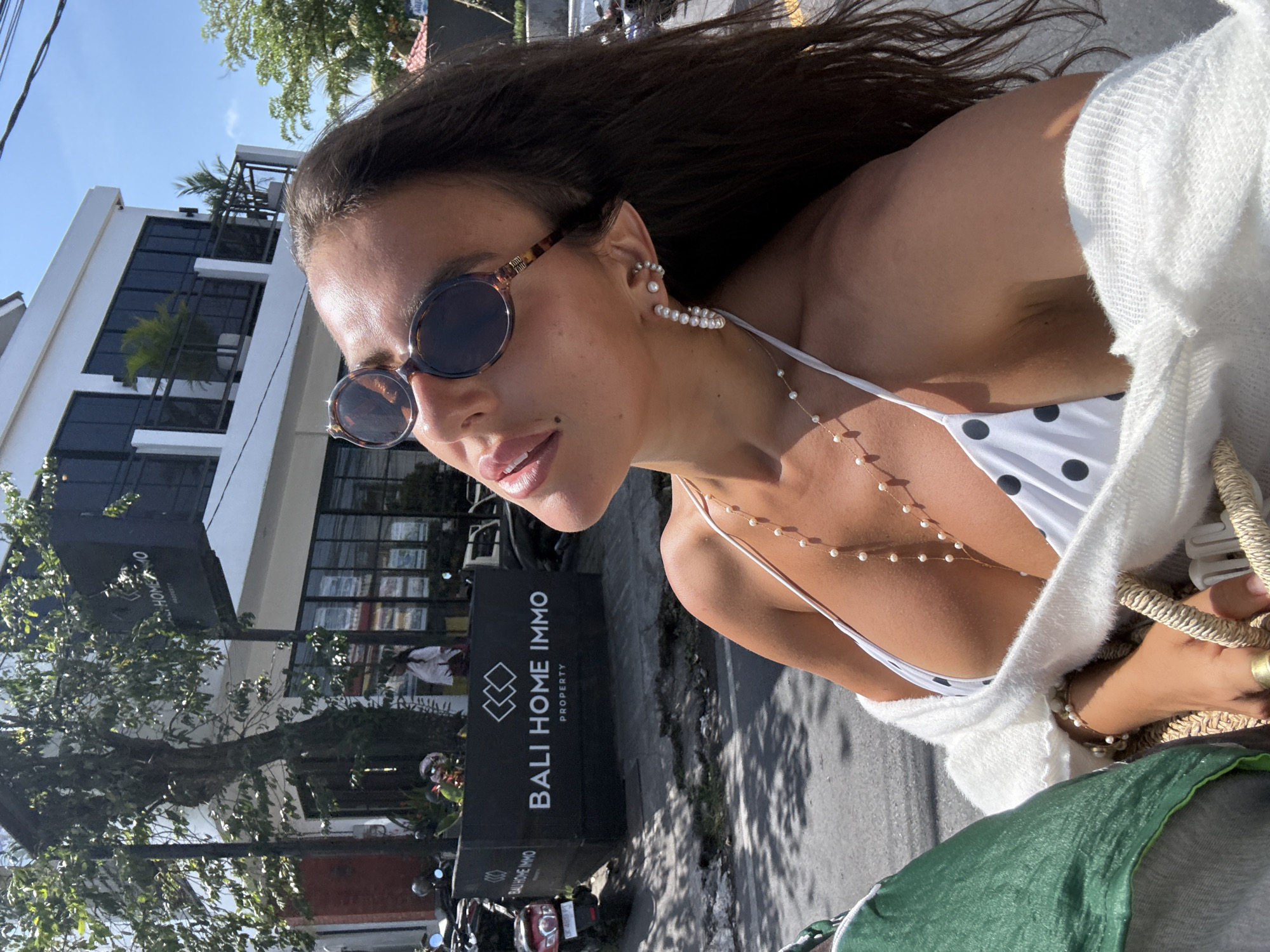 In Kuta Selatan, Indonesia, a woman is photographed outdoors, likely during the daytime. She is seen from the chest up, her head tilted back and eyes closed, with the sun on her face. She is wearing sunglasses with tortoiseshell frames, pearl earrings, and a white bikini with black polka dots, adorned with a delicate pearl necklace. A white, open-knit cardigan is draped over her shoulders. In the background, a white building with multiple windows and balconies stands under a clear blue sky, with lush green trees and foliage visible. To the left, a dark sign with white text reads "BALI HOME IMMO PROPERTY". Several motorcycles are parked on the pavement in front of the building, and a woman in a white shirt can be seen walking near the sign. The overall scene suggests a casual, sunny day in a tropical urban environment.