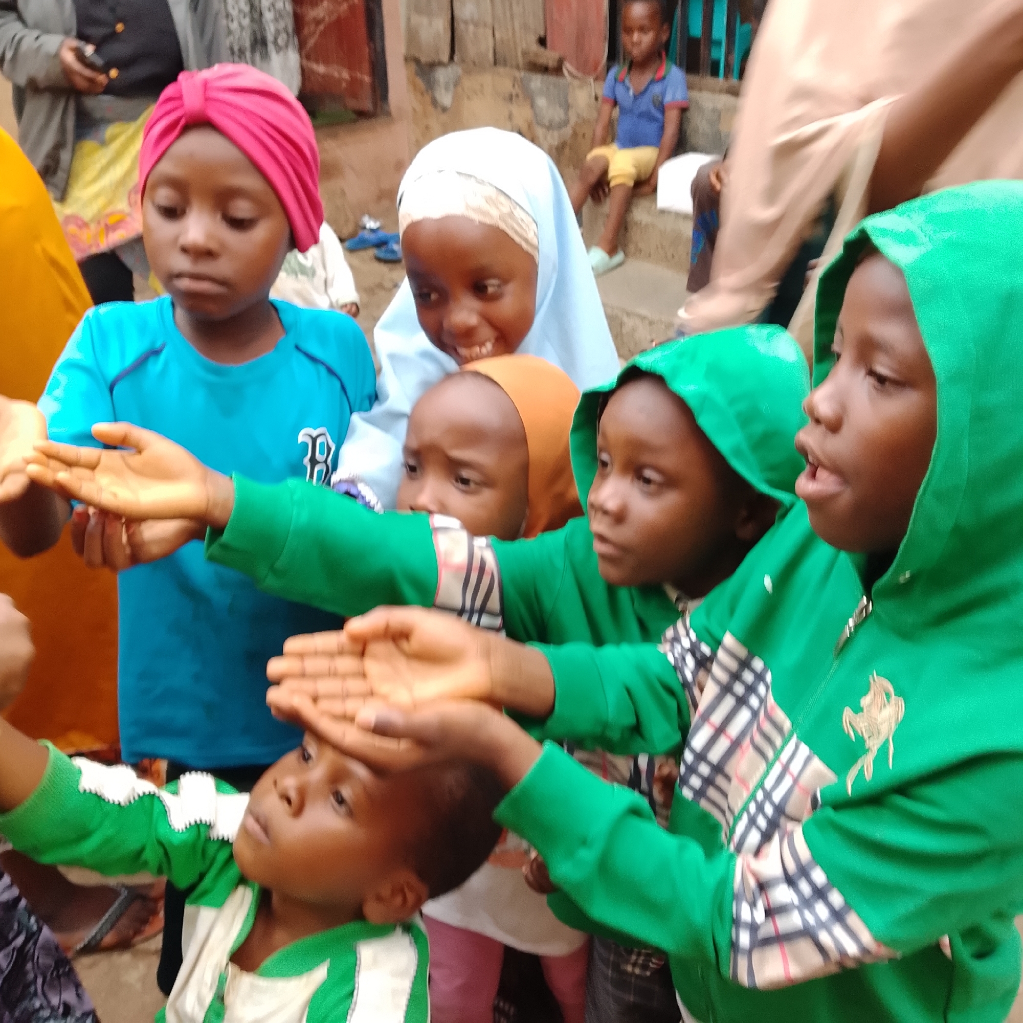 The image shows a group of children, some of whom are wearing headscarves and hoodies. In the foreground, a child in a bright green hoodie with plaid accents is reaching out their hands, with another child's hands touching their forehead. Another child to the left, wearing a blue shirt and a pink headscarf, is looking down. Behind them, a child in a light blue hijab is smiling. To the right, a young child in a blue shirt and yellow shorts is sitting on steps. The scene appears to be outdoors, with a rustic building in the background. The children's expressions suggest engagement in an activity or interaction. The time of day and weather are not explicitly discernible, but the general lighting suggests it might be daytime. No visible text is present in the image.