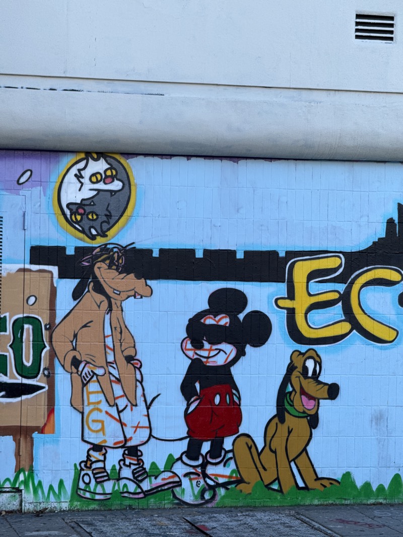 A vibrant mural decorates a wall in Los Angeles, United States. The mural depicts Goofy, Mickey Mouse, and Pluto stylized in a hip-hop aesthetic. Goofy is wearing sneakers, shorts, a t-shirt, and a jacket. Mickey Mouse is wearing sneakers and his classic attire. Pluto is laying on the ground to the right of Mickey Mouse. To the left of Goofy is a signpost with the letters 'GO' visible, and above Goofy and Mickey are two cats in a Ying and Yang position. A yellow 'EC' can be seen behind Pluto. The wall is pale blue with the exception of where the painting is present. The wall is constructed out of tiles. Above the painting is a white wall with an air vent.