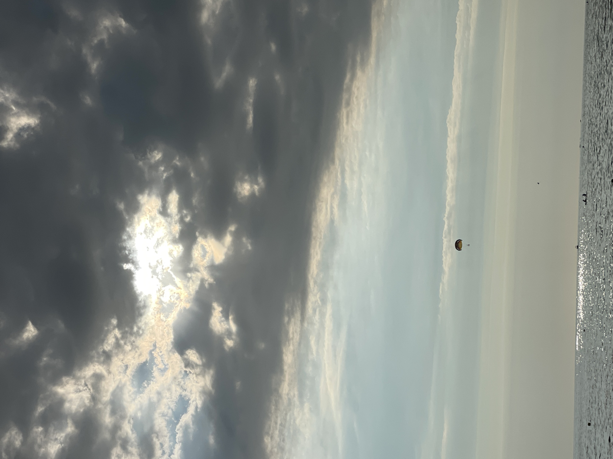 This image depicts a view of the Black Sea, likely near Sochi, Russia, characterized by a dramatic sky and a body of water. Dark, heavy clouds dominate the upper portion of the frame, interspersed with areas where the sun breaks through, casting rays of light. A single parasailer, with a colorful parachute, is visible in the sky, suspended above the water. The sea itself is calm, reflecting the subdued light of the sky, with small waves lapping at the shore. A few dark shapes, potentially buoys or small boats, are scattered across the water's surface. The overall atmosphere is moody and atmospheric, suggesting either an approaching storm or the aftermath of one.