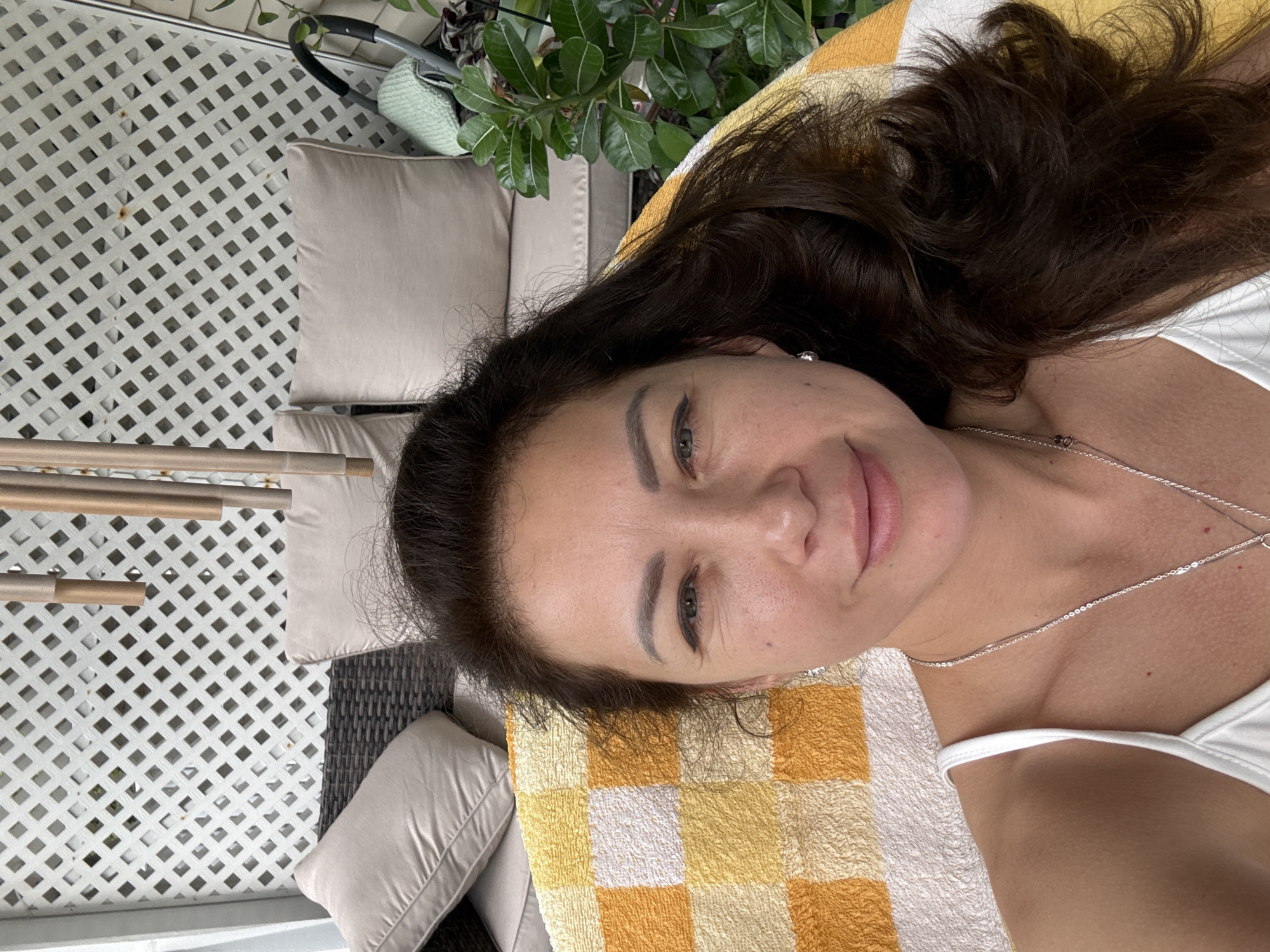 This is a close-up, overhead, selfie-style photograph taken outdoors on what appears to be a sunny day, likely in the afternoon. The subject is a woman with dark brown hair, lying down on a yellow and white checkered towel. She is wearing a white top and a silver necklace. Her face is turned towards the camera, with one eye visible and a slight smile. Behind her, a white lattice structure is visible, suggesting an outdoor patio or deck. Cushions from outdoor furniture are also visible, along with a potted plant with lush green leaves. The overall scene suggests a relaxed and leisurely moment, possibly at a resort or a home with outdoor living space. No text is visible in the image.