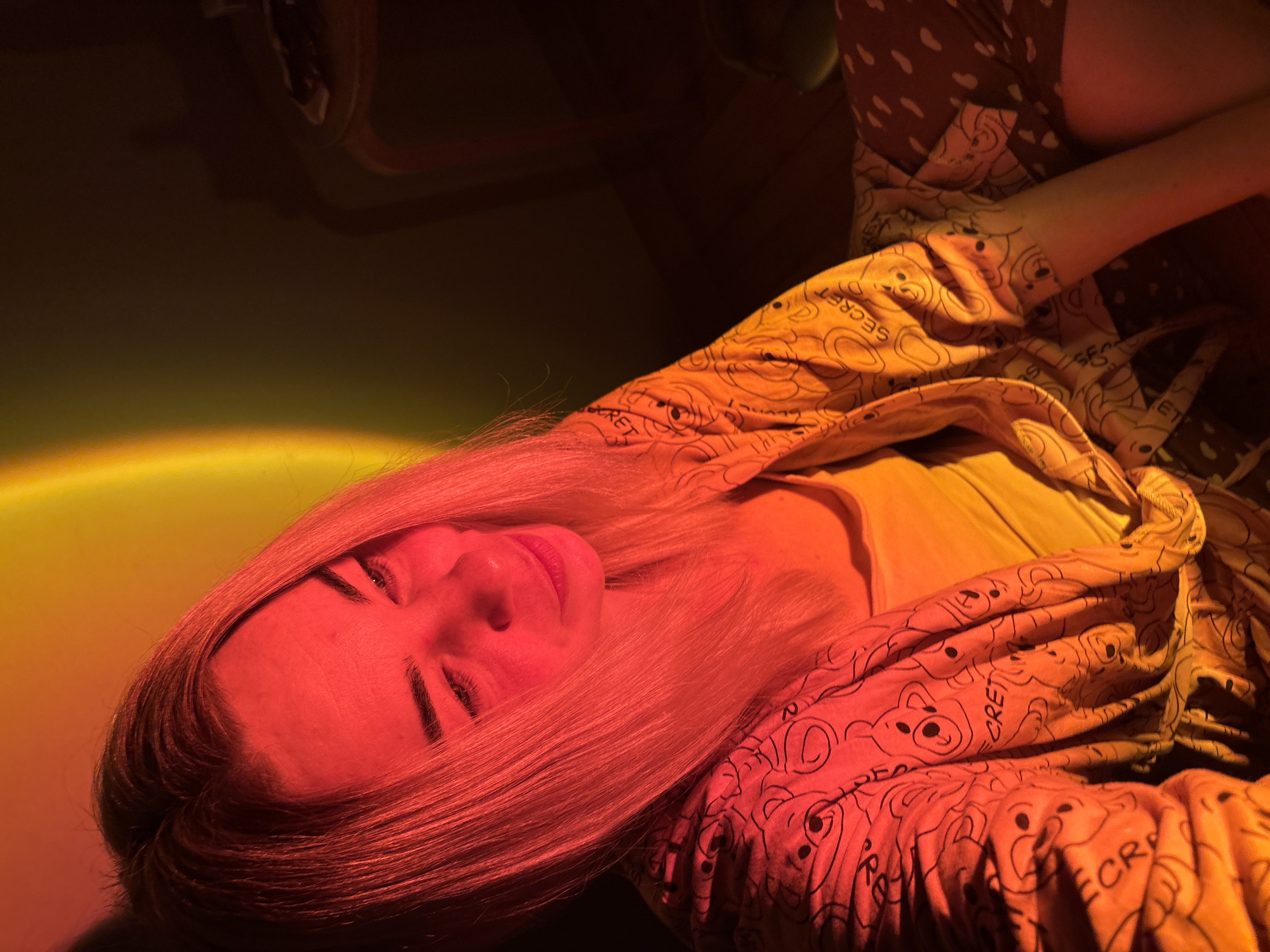 A young Caucasian woman with blonde hair, wearing a yellow tank top and a printed robe, is sitting on the floor indoors. The room is dimly lit, with a warm red and orange light cast by a projection, likely from a sunset lamp, on the wall and illuminating the woman. Her gaze is directed downwards and to her right, with a slight smile on her face. The robe she is wearing has a pattern of cartoon bears and the word "SECRET" repeated. The floor appears to be wooden. In the background, a dark wall is visible, and a small, dark piece of furniture with a curved leg is to the right. There is no indication of the woman's location within the city of Sinyavino, Russia; the image provides no geographical context for the city.