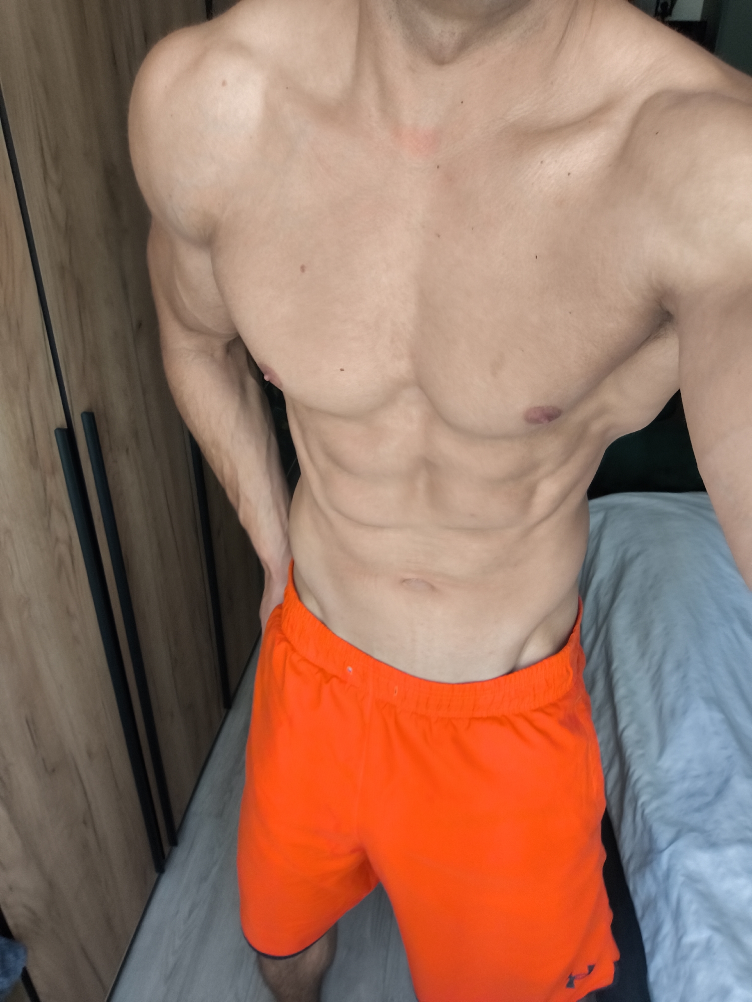 An adult male with a muscular, lean physique is depicted from the upper chest to mid-thigh, standing in an indoor setting. He has visible muscle definition in his chest, shoulders, and a prominent six-pack abdomen. His skin is light-toned, with several small dark moles scattered across his torso. A faint reddish mark is present near his left clavicle, and his right nipple area shows a slight reddish-purple discoloration. A small scar or mark is visible above his navel. He is wearing bright orange athletic shorts with a dark-colored trim along the bottom edges; a black "Under Armour" logo is partially visible on the lower right leg of the shorts. His left hand is placed on his left hip.
The background on the left side of the frame features a wooden wardrobe or cabinet with a light-to-medium wood grain pattern, equipped with three vertical dark-colored (likely black) handles or trim elements. The floor directly beneath is a light-colored wood laminate or similar material. On the right side, a bed with light blue or grey bedding is visible, with a darker area (possibly a wall or headboard) behind it. The overall environment appears to be a bedroom or dressing area.
