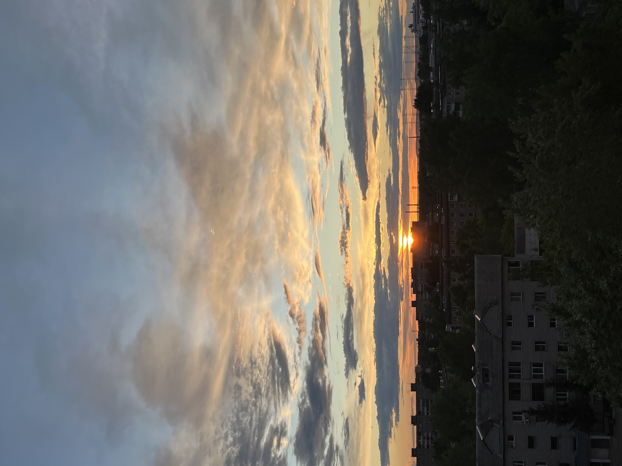 The image captures a sunset in an urban setting. The sky is filled with clouds illuminated by the setting sun, creating a mix of golden, orange, and blue hues. Below the sky is a cityscape with buildings and trees. The sun is positioned low on the horizon. The exact location is undetermined without additional context.