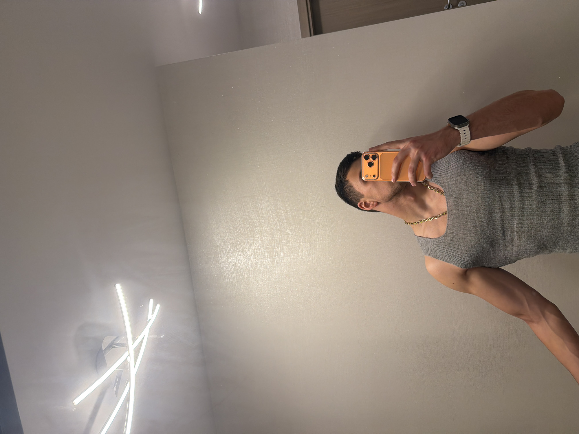 An upward-oriented mirror selfie captures a man's partial reflection against a light-toned, subtly textured wall. The image is rotated 90 degrees clockwise, presenting the man as if lying on his back with his head to the right.
The subject is a man with dark hair, visible from the right side of his head, including his ear and jawline. He is wearing a gray, ribbed sleeveless top or tank top, and a prominent gold-colored chain necklace is around his neck. On his left wrist, a smartwatch with a light-colored band is visible. He holds an orange or gold-colored smartphone in his right hand, covering the central part of his face. The phone clearly shows a triple camera setup.
The background, seen through the mirror, is a plain, light-colored wall, possibly cream or light gray, with a fine, shimmery or textured finish that catches the light. In the lower left portion of the reflection, a modern, abstract light fixture with multiple white, glowing bar-like elements is prominently featured. The lighting appears to be artificial and bright, suggesting an indoor setting. There are no overt location cues beyond an interior room, and no discernible text is visible in the image.