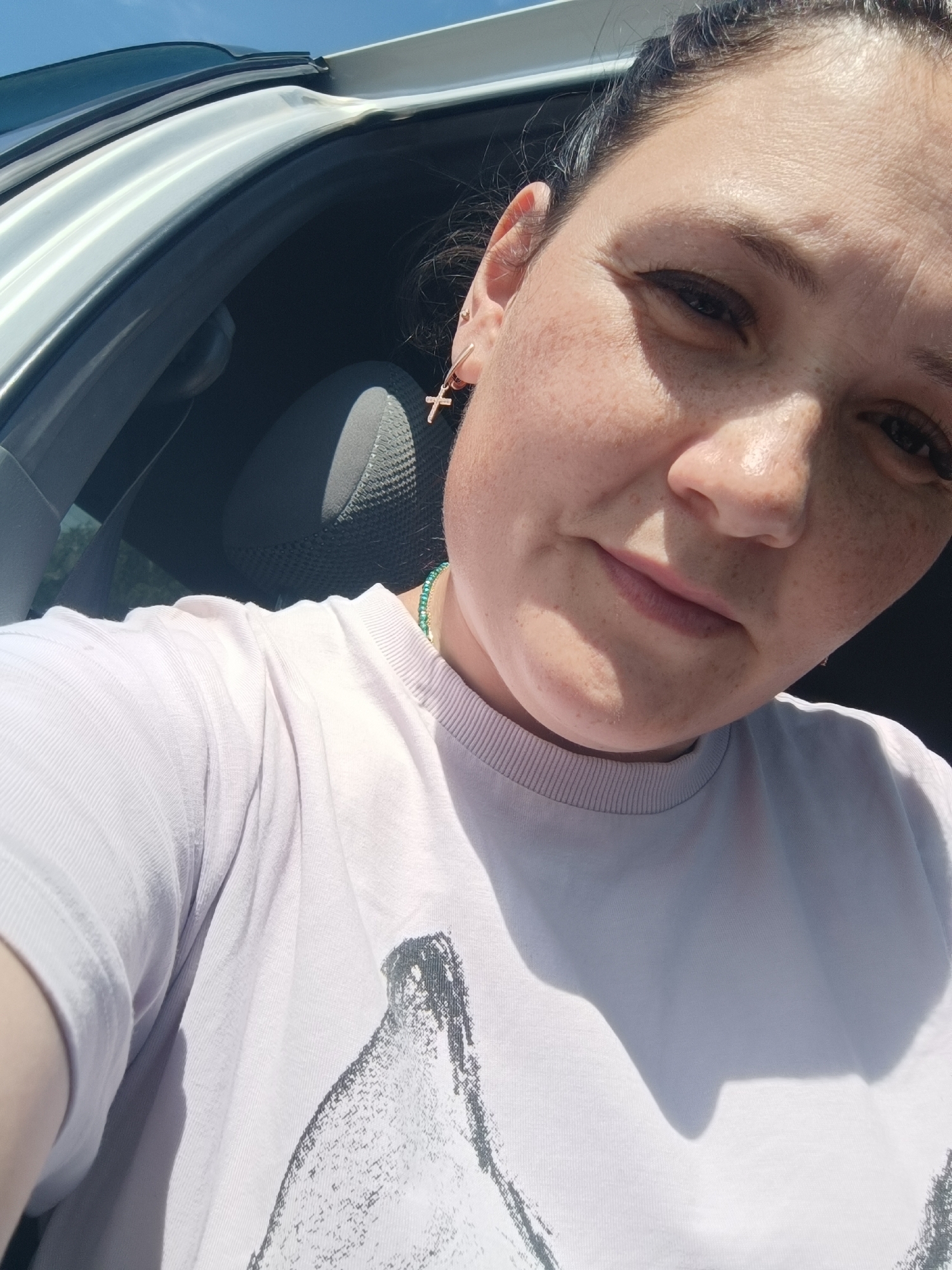 The image is a selfie taken by a woman inside a vehicle, likely in Chaikovsky, Russia. The woman has fair skin with freckles and is wearing a light-colored t-shirt with a graphic design. She has a cross-shaped earring in her left ear and is wearing a beaded necklace. The background shows the interior of the car, including a seat and a window with a view of the sky. The lighting suggests it is daytime, and the sun is casting shadows on the woman and her clothing.
