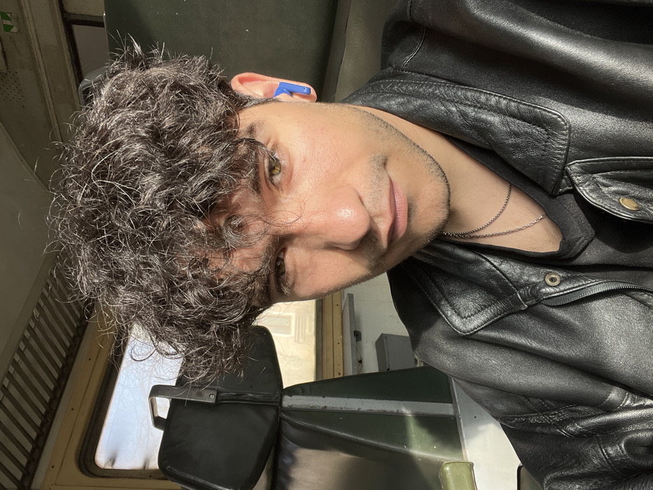 A young adult male is depicted in a close-up, waist-up portrait, seated inside what appears to be a passenger compartment of a train or bus in Todoretsite, Bulgaria. The male subject has dark, curly hair and light stubble, with brown or hazel eyes, and looks directly at the camera with a neutral expression. He wears a black leather jacket over a dark t-shirt, accessorized with a thin silver chain necklace. A single blue wireless earbud is visible in his right ear. Strong natural light, consistent with sunlight, illuminates the left side of his face. In the background, dark-colored, possibly green or black, upholstered seats are partially visible. A black rectangular bag with a silver handle rests on the seat behind him. Through a window located to the upper left and behind the subject, a blurred view of an exterior environment, suggesting buildings or structures, is discernible. The vehicle's interior features include ribbed paneling on the ceiling.