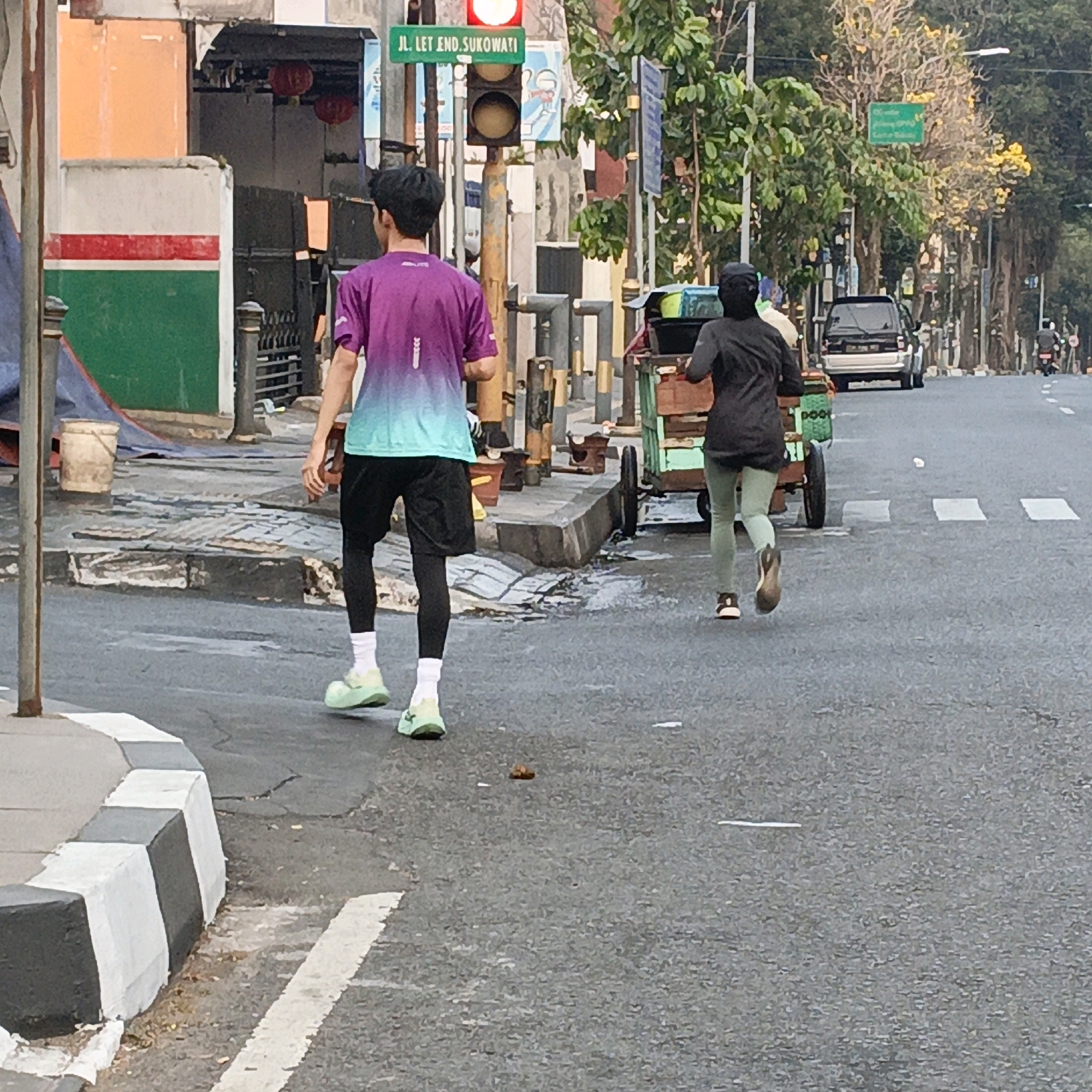 A young man wearing a purple and turquoise ombre t-shirt, black shorts, black compression pants, white socks, and light green athletic shoes is walking away from the camera on a paved road. He is on the right side of the road, walking alongside a cart with green and brown components being pulled by a person behind him, who is partially visible and wearing a dark top and light green pants.
The scene appears to be in an urban environment in Salatiga, Indonesia. Behind the first man is a street with a traffic light and a green street sign that reads "JL. LET END. SUKOWATI". There are buildings to the left of the street with red lanterns visible, and trees line the right side of the road. The road surface shows signs of wear, with some cracks and white painted markings. The day appears to be overcast.