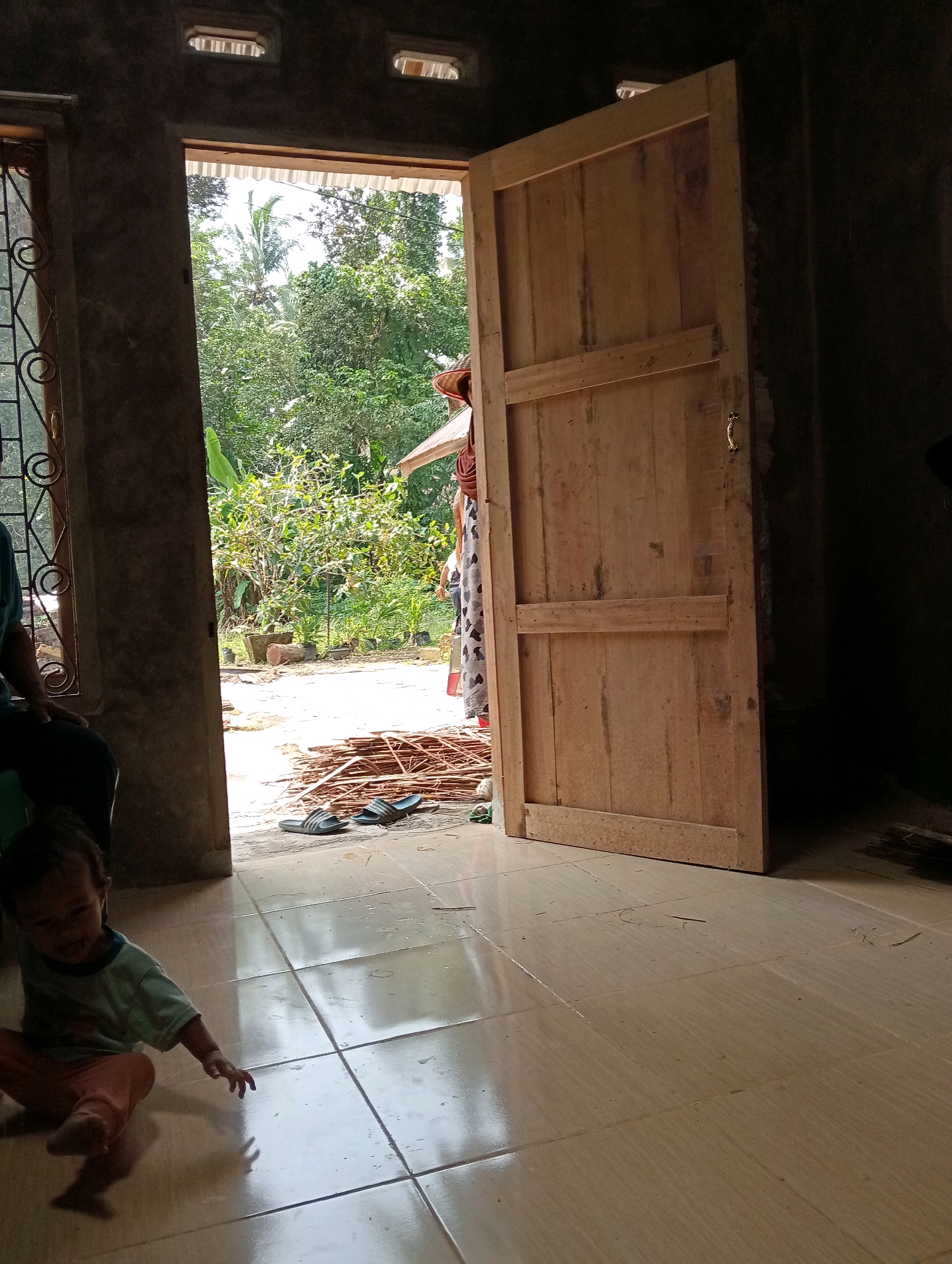 The image depicts an interior scene looking out through an open doorway at a rural outdoor environment. The foreground is dominated by a tiled floor. A young child, wearing a light blue t-shirt and orange pants, is seated on the floor on the left side of the image, facing right and reaching out with their right hand. To the right of the child, a pair of black and white striped flip-flops are placed on the floor near the doorway.
The doorway itself is wide and made of unpainted wood, with a simple horizontal brace across the lower half and a gold-colored latch. Beyond the doorway, the scene opens to a sunlit outdoor area with lush green vegetation, including trees and possibly banana leaves. A pathway or ground surface is visible leading away from the house. In the middle ground, to the right of the doorway, a person wearing a patterned dress and a conical hat is partially visible, looking outwards. A stack of dried palm fronds or similar material is piled on the ground near the doorway. The interior walls are dark and appear to be made of concrete. Two rectangular vents are visible high on the interior wall above the doorway. The overall impression is of a simple, rural dwelling in Campago Barat, Indonesia, with the interior space connecting to the natural environment outside.