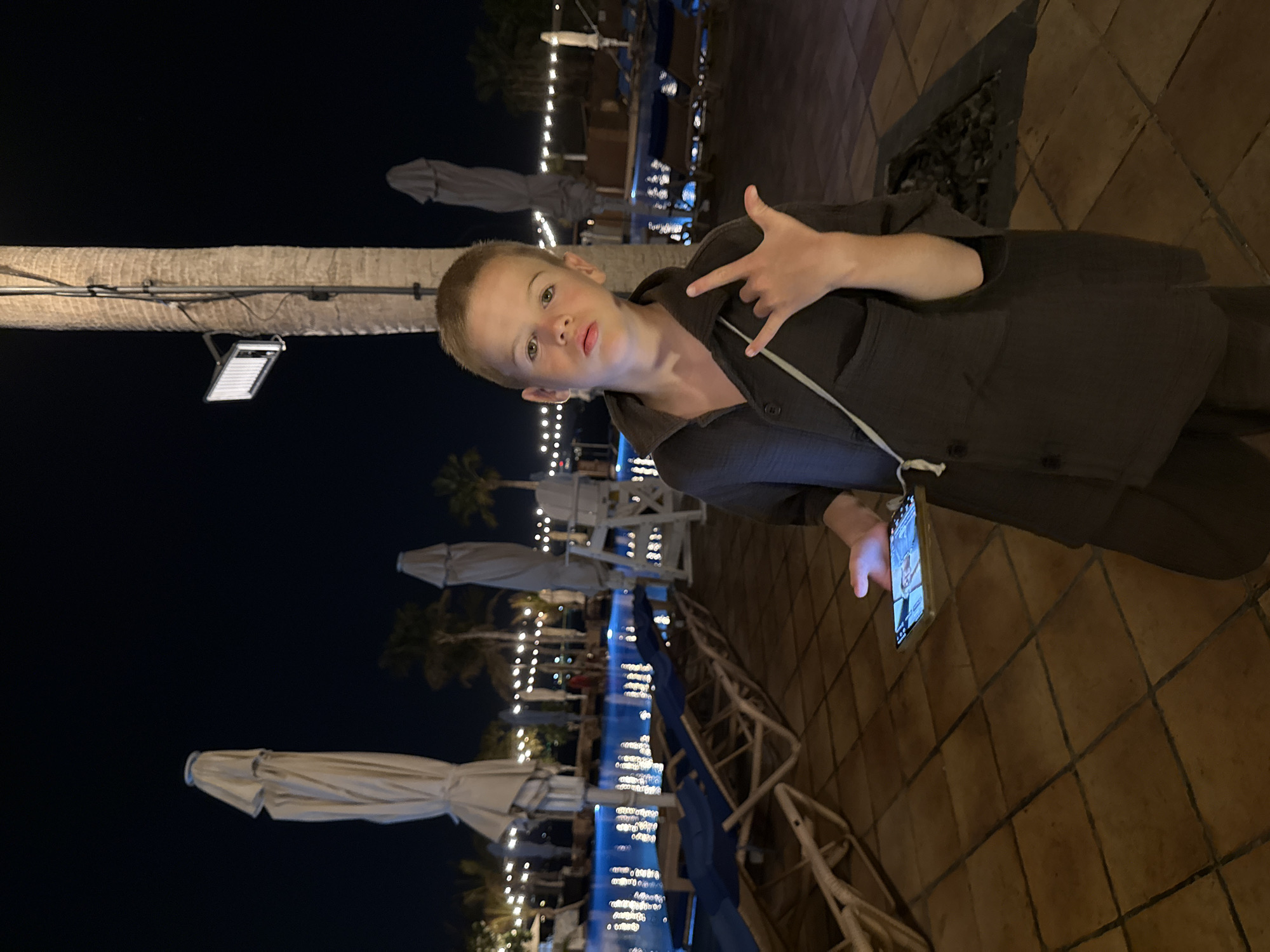 A young boy is posing for a photo at night. He is wearing a dark-colored jacket and has his right hand raised in a rock-on gesture. He is looking towards the camera with a playful expression. The boy is standing on a tiled surface, and behind him, a swimming pool with blue lights is visible, along with palm trees and string lights. Umbrellas and lounge chairs are set up around the pool area. A smartphone is being held by someone outside the frame, displaying a video call with two other people. The overall setting appears to be a resort or hotel in Dubai, UAE, at night, with warm lighting from the illuminated pool and decorative lights. The weather appears to be clear.