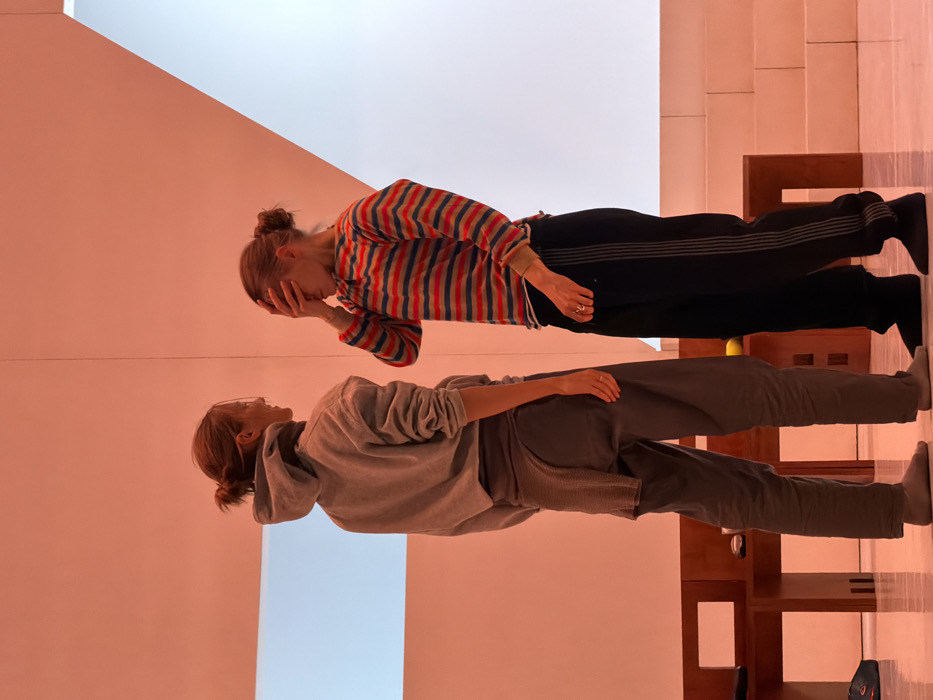 Two individuals are positioned in a modern, minimalist indoor environment bathed in soft, light orange ambient lighting. The setting, characterized by stark white and light orange walls, angular architectural elements, and minimalist furnishings, suggests a contemporary art exhibition space, performance venue, or studio in Paris, France, likely a cultural institution such as the Fondation Louis Vuitton.
On the left, an individual, appearing to be female, stands facing right, partially obscured from a rear-side view. She wears a light grey hooded sweatshirt, dark grey loose-fitting trousers, and light grey socks. A piece of grey, subtly patterned fabric is wrapped around her waist. Her light brown hair is pulled back into a bun, and she is looking towards the other person.
On the right, another individual, also appearing female, stands facing left, with her body angled slightly. She wears a horizontally striped long-sleeved shirt featuring red, orange, blue, and white stripes, black track pants with two grey vertical stripes down the side, and black socks. Her right hand covers her face, obscuring her eyes and forehead. A metallic ring is visible on her left ring finger. Her light brown hair is tied in a bun.
In the mid-ground, between the two individuals, two low, minimalist wooden benches or tables are present. A small yellow object and a dark, indistinct item rest on one of the benches. The background includes a rectangular white panel inset into the left wall and a large, angular white structure, resembling a sloped wall or a series of steps, on the right. The floor is light-colored and subtly reflective.