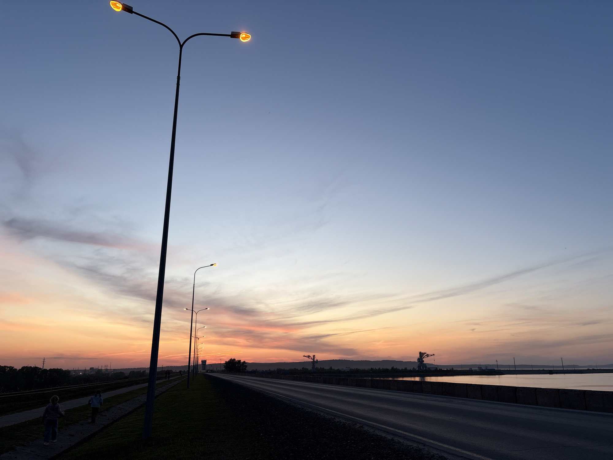 The image depicts a view of the city of Chaikovsky, Russia, at sunrise or sunset. A long, straight road with a low concrete barrier on the right stretches into the distance. To the left of the road, a row of streetlights illuminate the scene. Two children are walking along a path beside a railway track. In the background, silhouetted industrial structures, possibly cranes, are visible against the horizon. The sky transitions from deep blue at the top to a gradient of orange and pink near the horizon. The scene is illuminated by the ambient light of the sky and the glow of the streetlights.