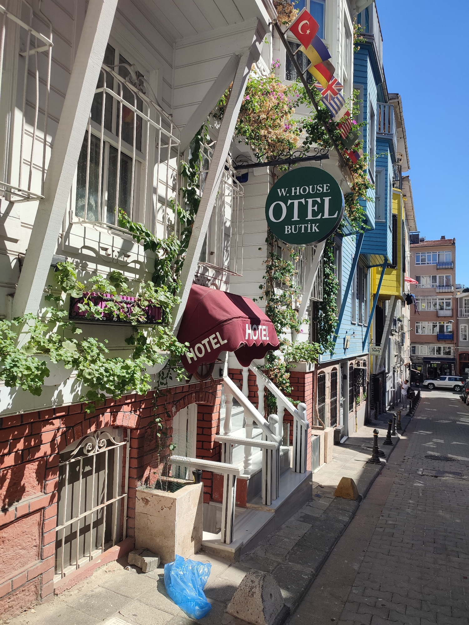 The image shows a street scene in the Kadıköy district of Istanbul, Turkey. In the foreground, a white building with red brick accents has a red awning that says "HOTEL". Above the awning, there are plants growing on the facade. A green sign reads "W. HOUSE OTEL BUTIK" with several international flags displayed above it. To the right of this building, there is a vibrant blue and yellow building, followed by more buildings of varying colors and heights. A narrow, paved street runs alongside the buildings, lined with bollards. The street appears to be a pedestrian walkway with some vehicular traffic further down. A person is visible in the distance, interacting with a vehicle. The sky is clear and blue.