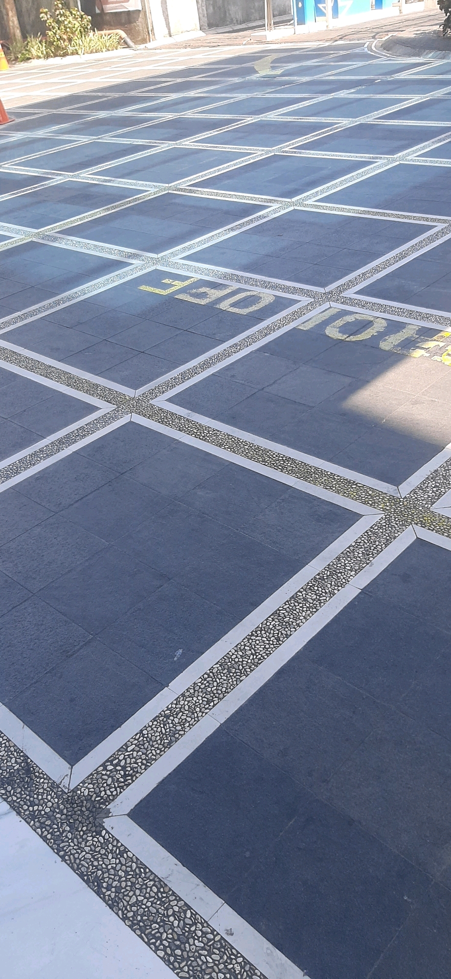 The image shows an outdoor paved area with a grid pattern of dark square tiles. The grid lines are formed by a combination of white borders and a band of small, light-colored pebbles. Yellow painted text is visible on some of the dark tiles, forming the words "TOLL" and "OFF". In the upper left corner, there is a sliver of a bright orange traffic cone and some green foliage. The background shows a wall and a building with blue accents. The perspective is from above, looking down at the patterned pavement. The scene appears to be a walkway or public space. Without additional information or context, the specific location within Tasikmalaya, Indonesia, cannot be determined from the image alone.